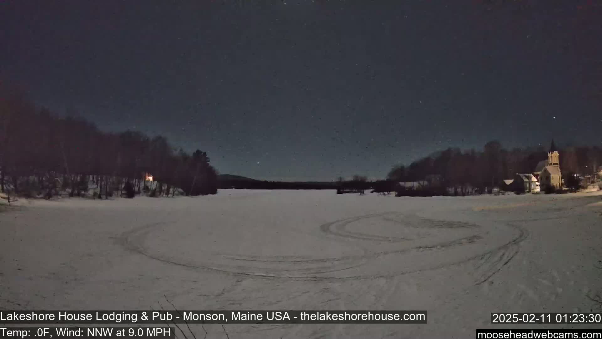A snow-covered, frozen lake at night under a starry sky, with a few buildings and trees visible in the distance, shows tire tracks in the snow.
