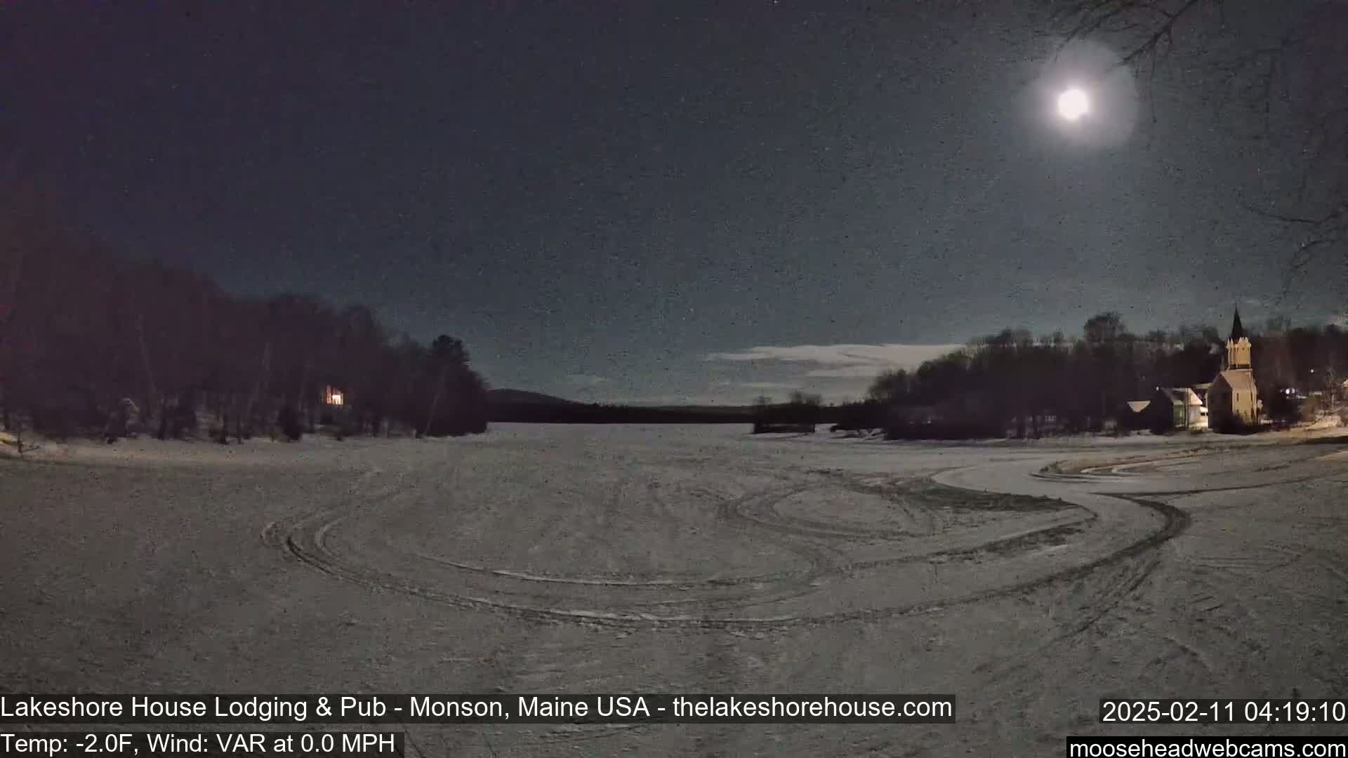 A bright moon illuminates a snow-covered frozen lake with tire tracks and a church in the background under a clear night sky.