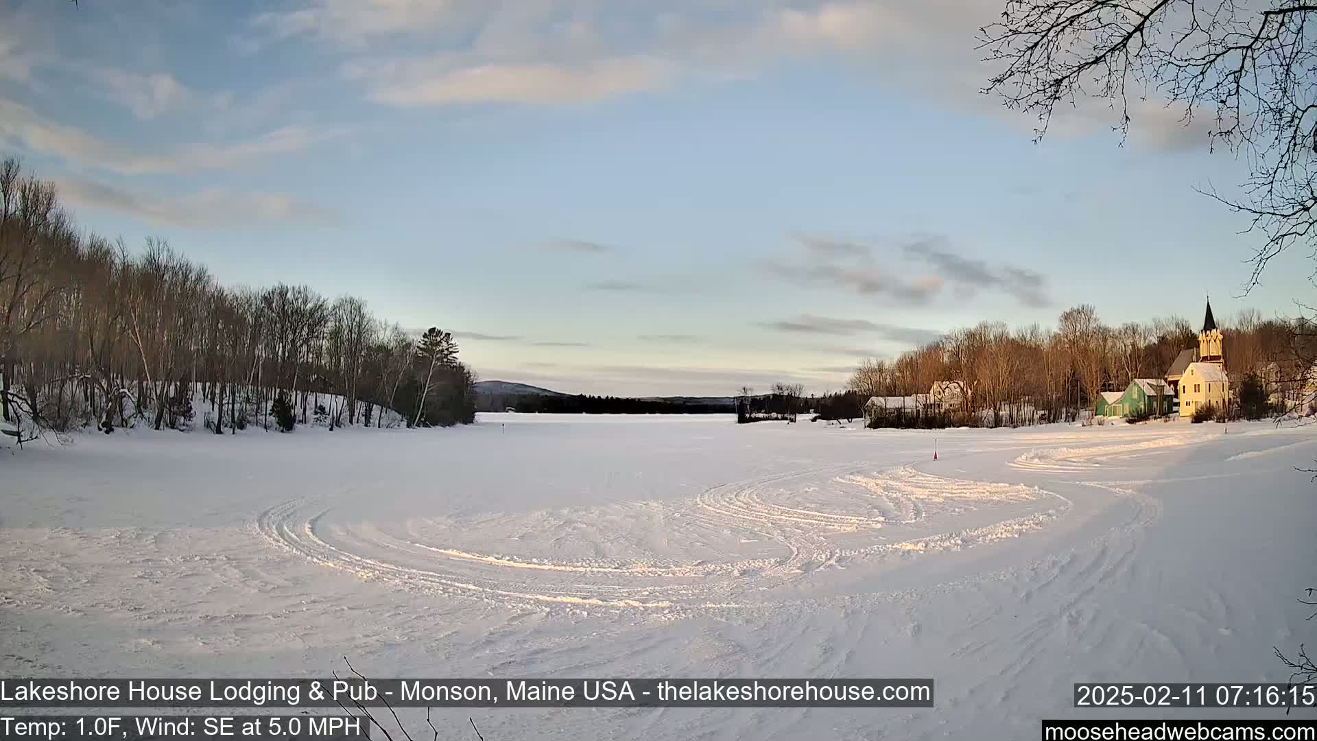 A snow-covered frozen lake with tire tracks, surrounded by leafless trees and a small village with buildings including a church steeple, under a partly cloudy sky.