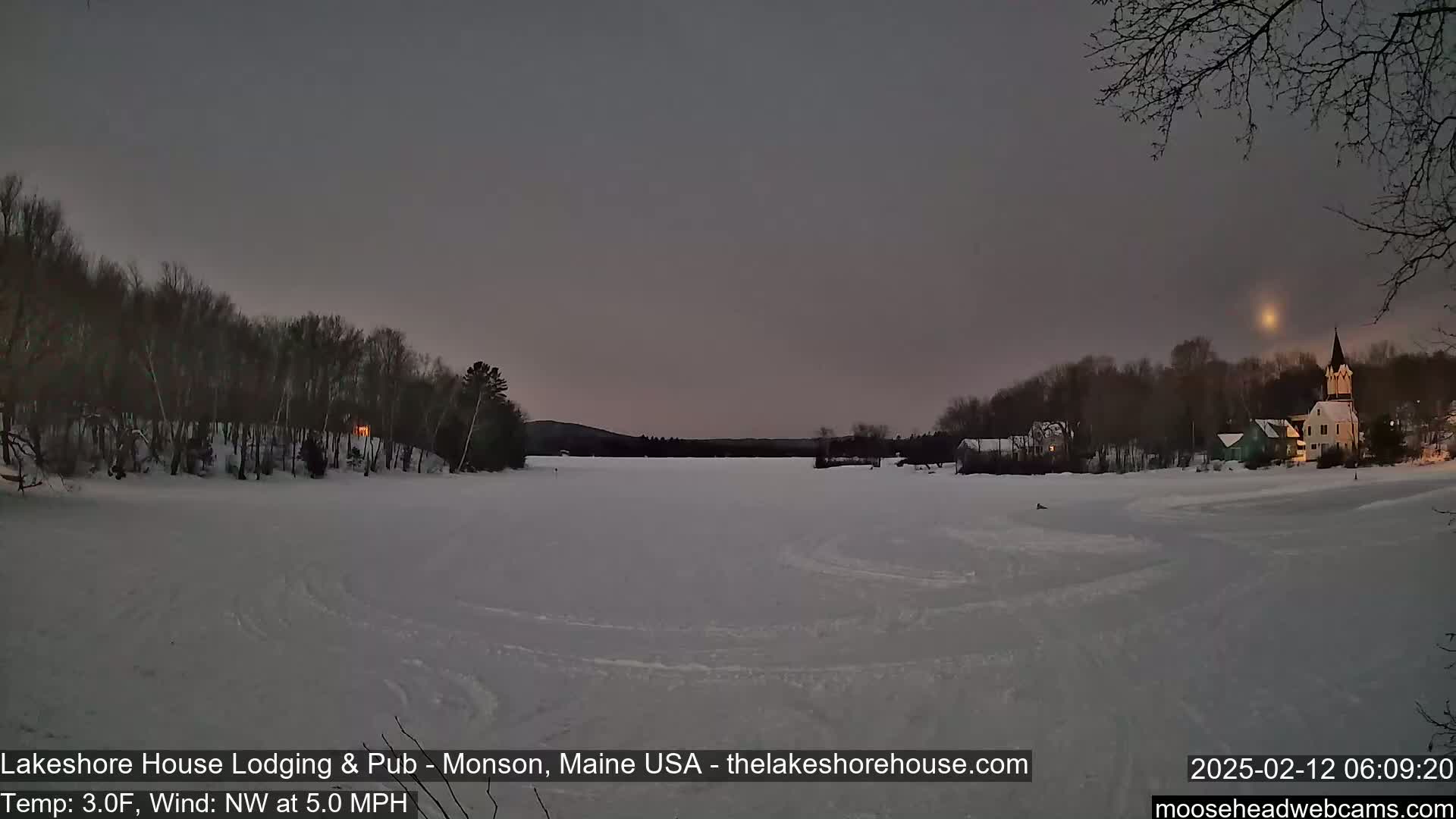 A frozen lake at twilight, under a dusky sky, is surrounded by snow-covered trees and buildings with lights visible in some windows.