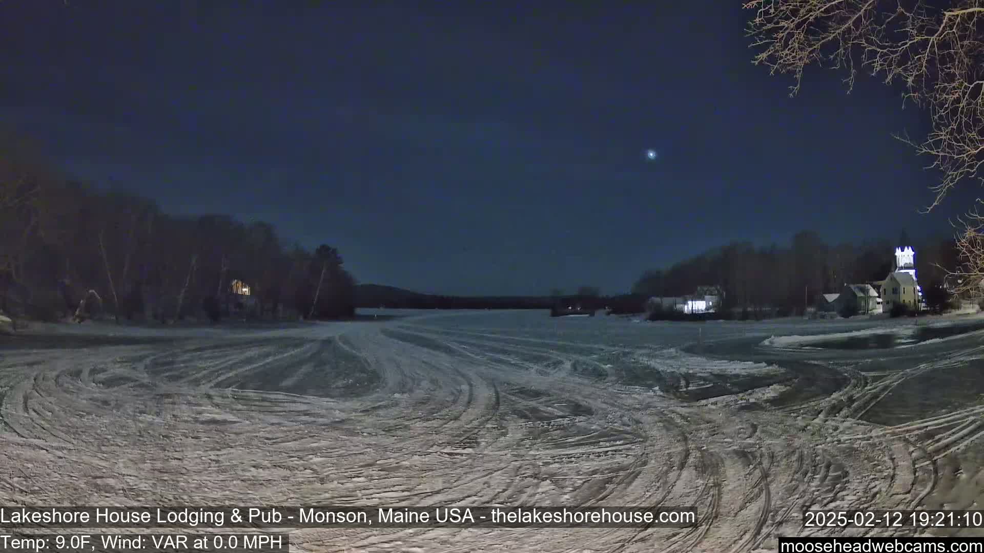 A frozen lake at night, under a dark sky with a single bright star, shows tire tracks across its surface and lit buildings on the far shore.