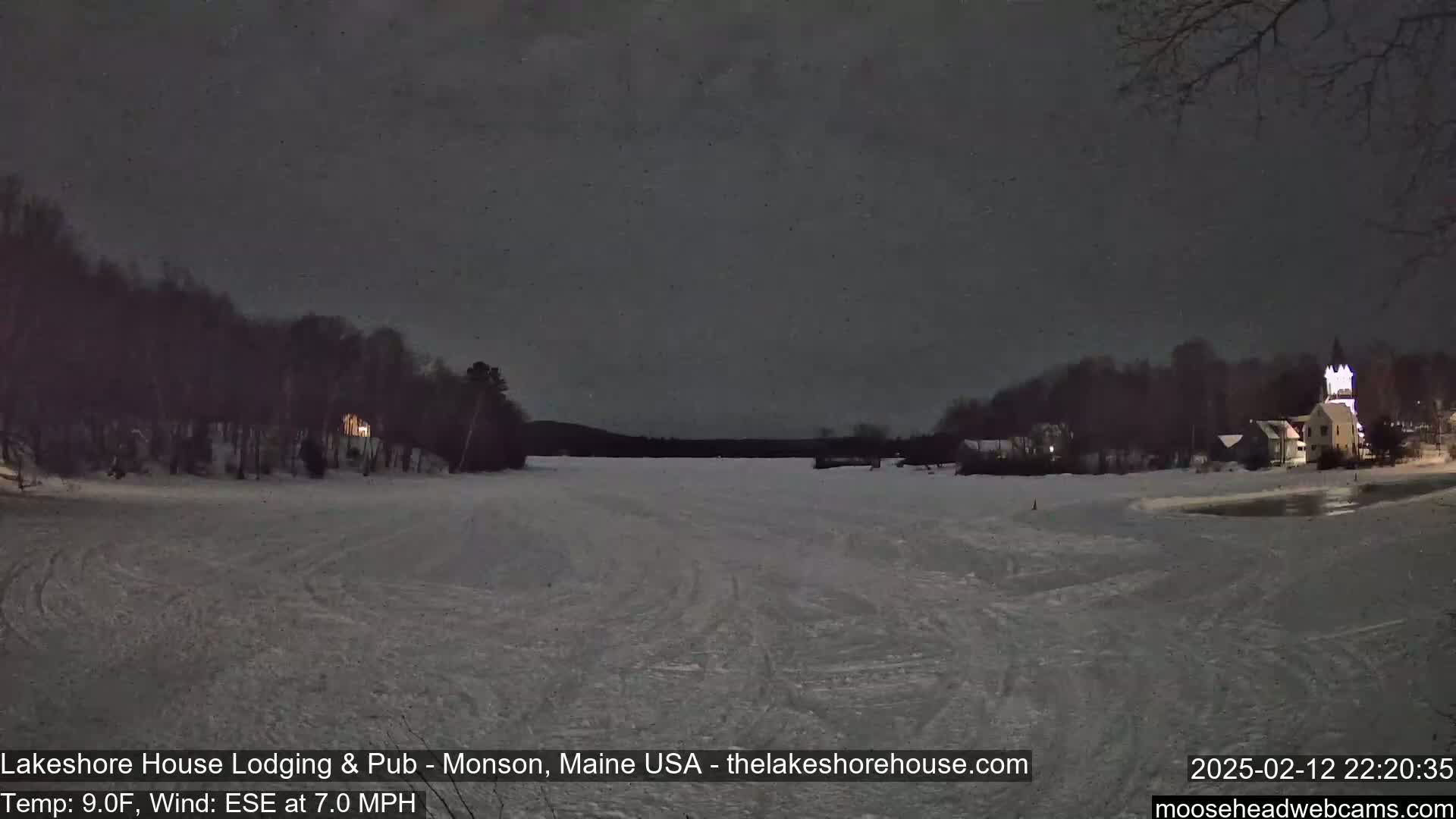 A snow-covered frozen lake at night, under a dark sky, is bordered by dark trees and illuminated buildings.