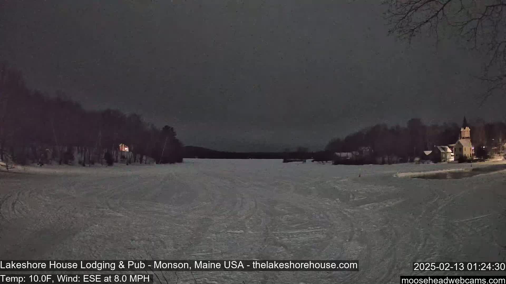 A snow-covered frozen lake is seen at night, with dark silhouettes of trees and buildings lining the shore under a dark sky.