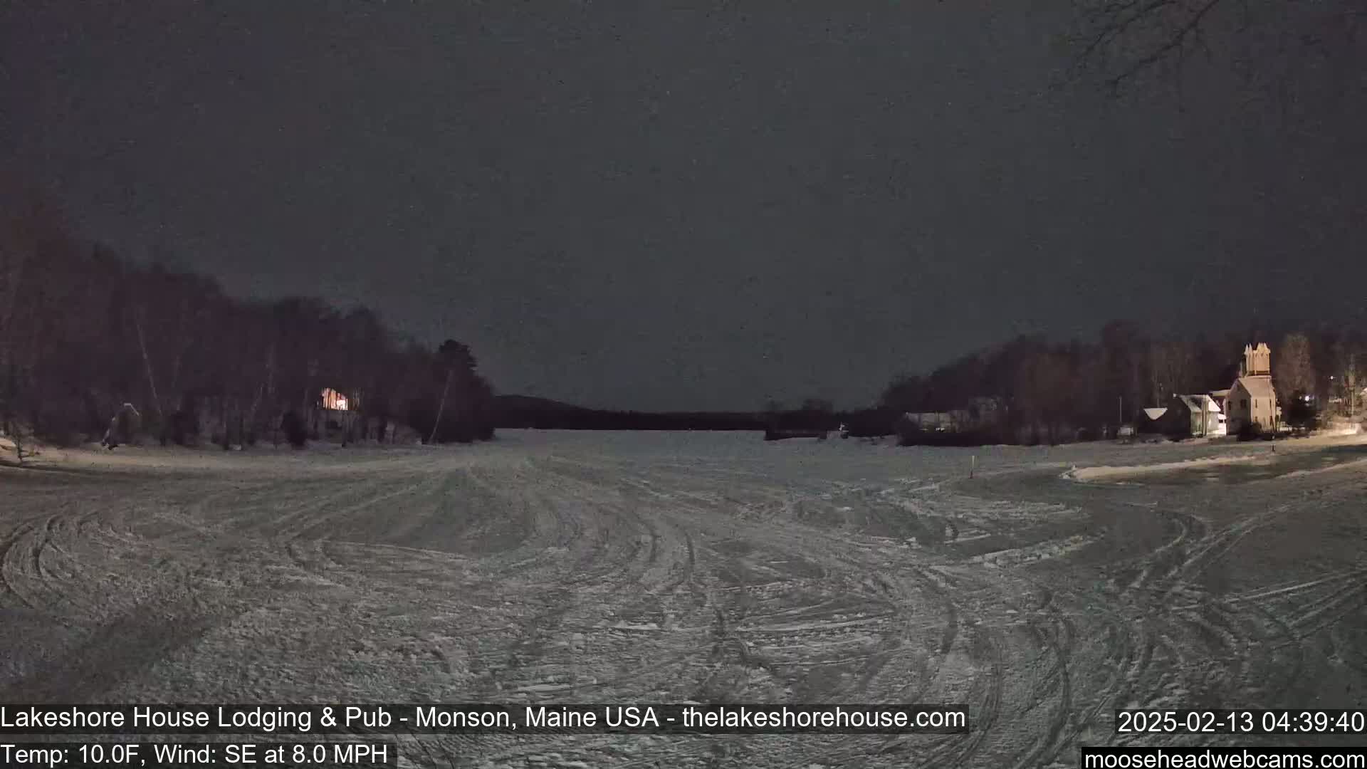 A nighttime view of a snow-covered frozen lake with trees and buildings on either side under a dark sky.