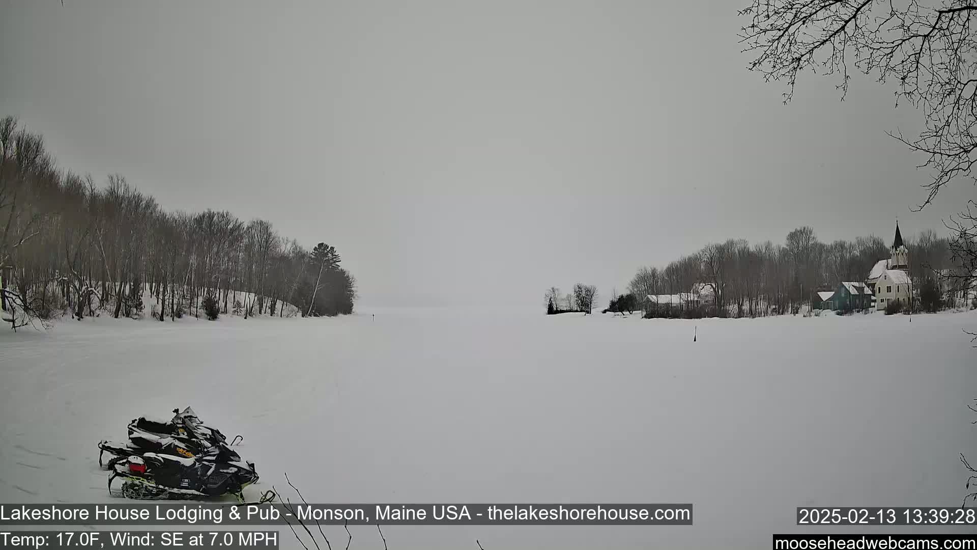 A snow-covered lake with a village and snowmobiles parked on the shore under an overcast sky.
