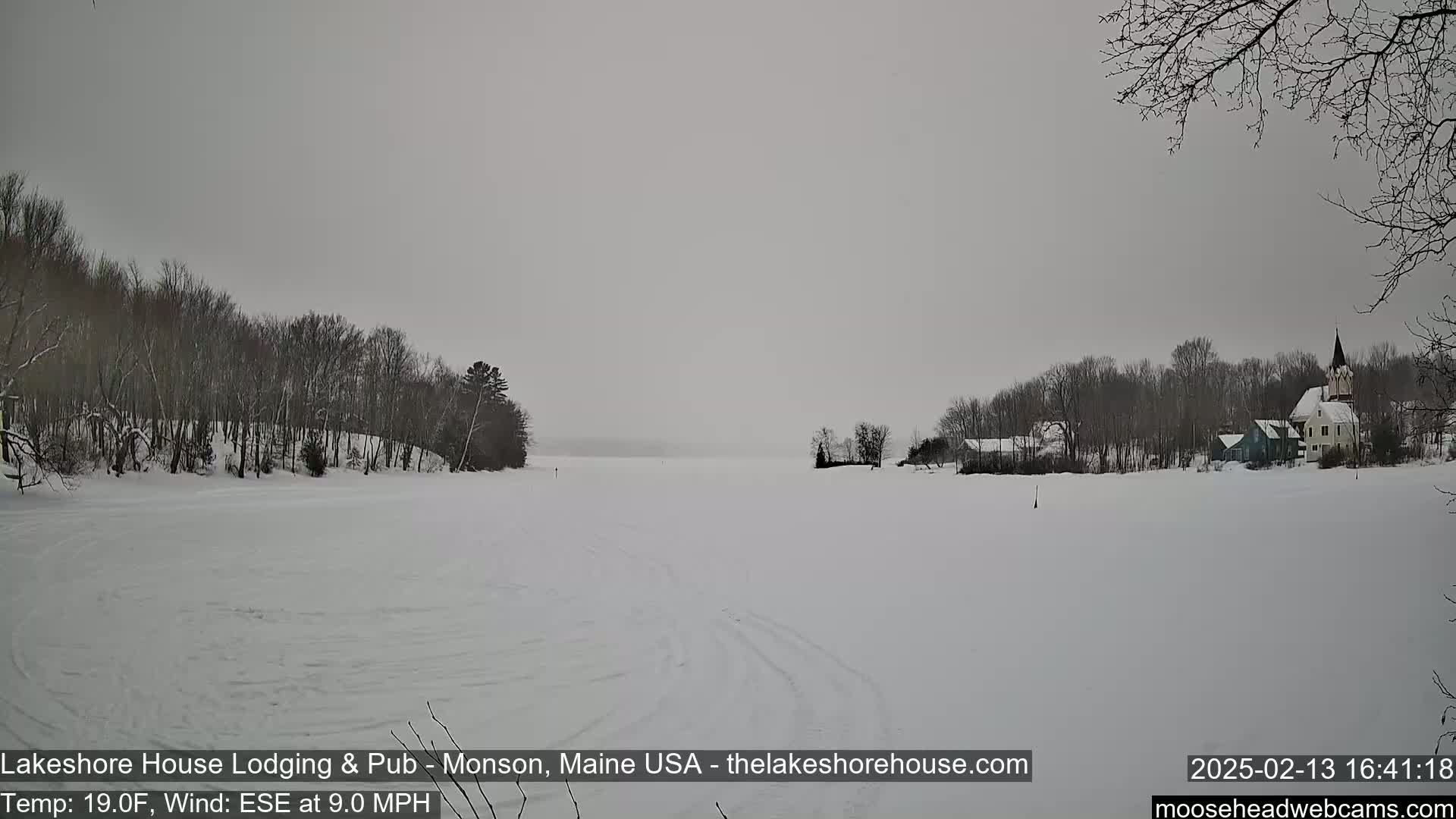 A snow-covered frozen lake stretches out before a line of snow-covered trees and houses, under an overcast sky.