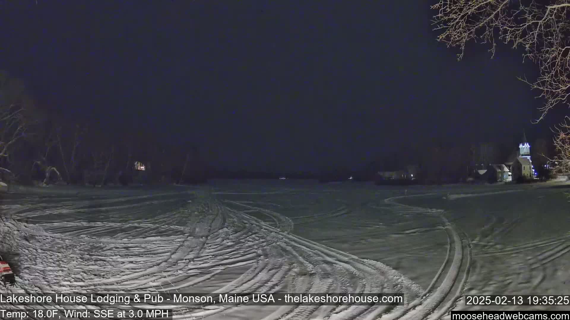 A snow-covered, flat expanse with tire tracks under a dark night sky shows faint lights from buildings in the distance.