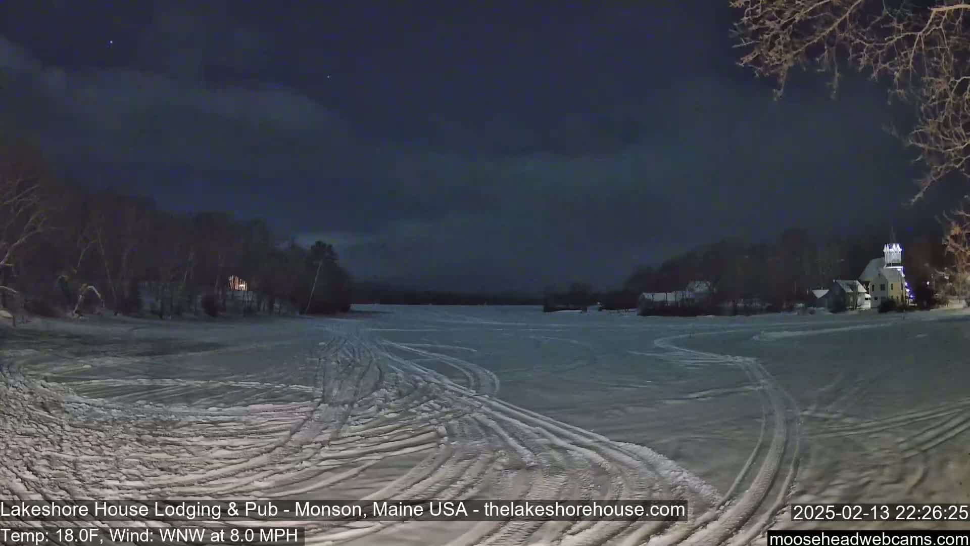 A frozen lake at night, under a dark, mostly clear sky, shows tire tracks across its surface, with buildings and trees visible on the far shore.
