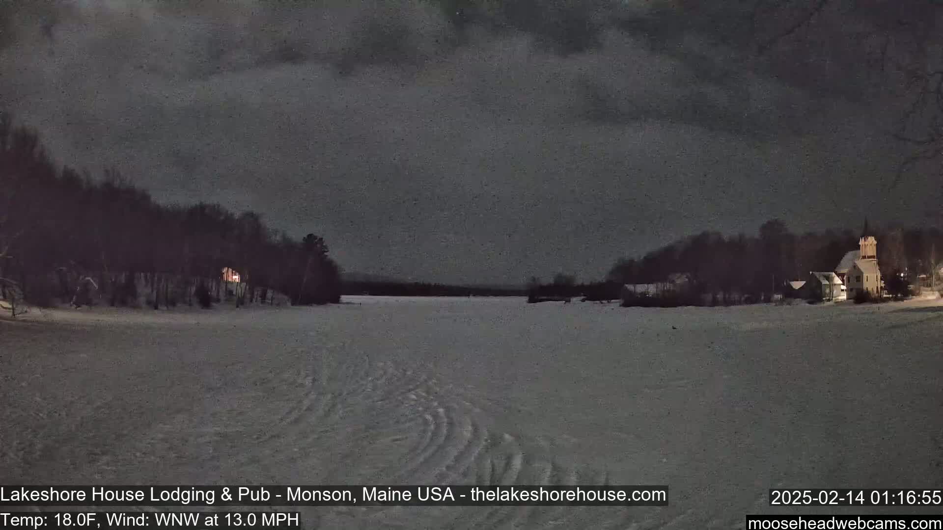 A nighttime view of a snow-covered lake, with dark trees lining the shores and buildings visible in the distance under a dark, overcast sky.