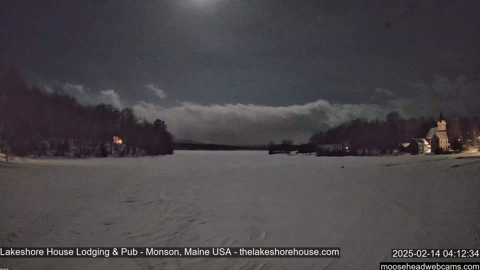 A snow-covered landscape at night, illuminated by a bright moon and the lights of buildings across a frozen lake under a partly cloudy sky.
