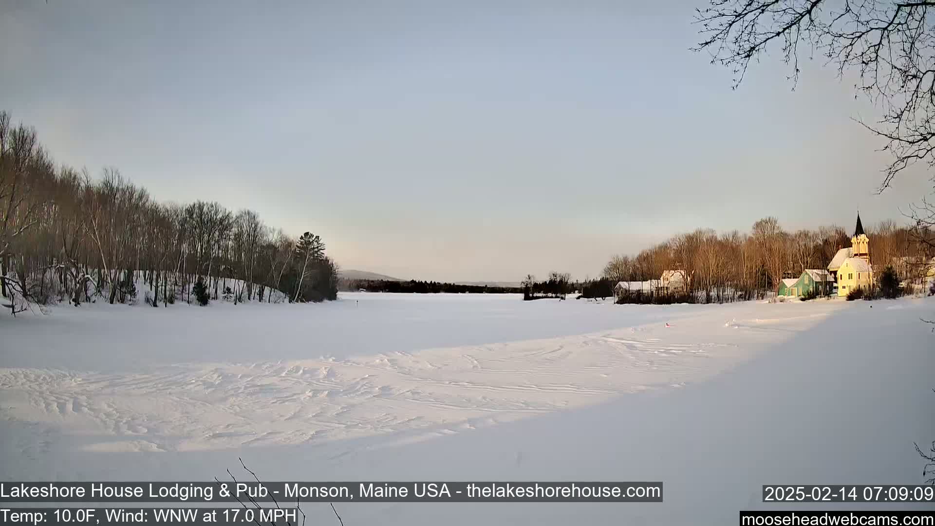 A snow-covered frozen lake stretches out before a line of houses and trees under a pale blue sky.