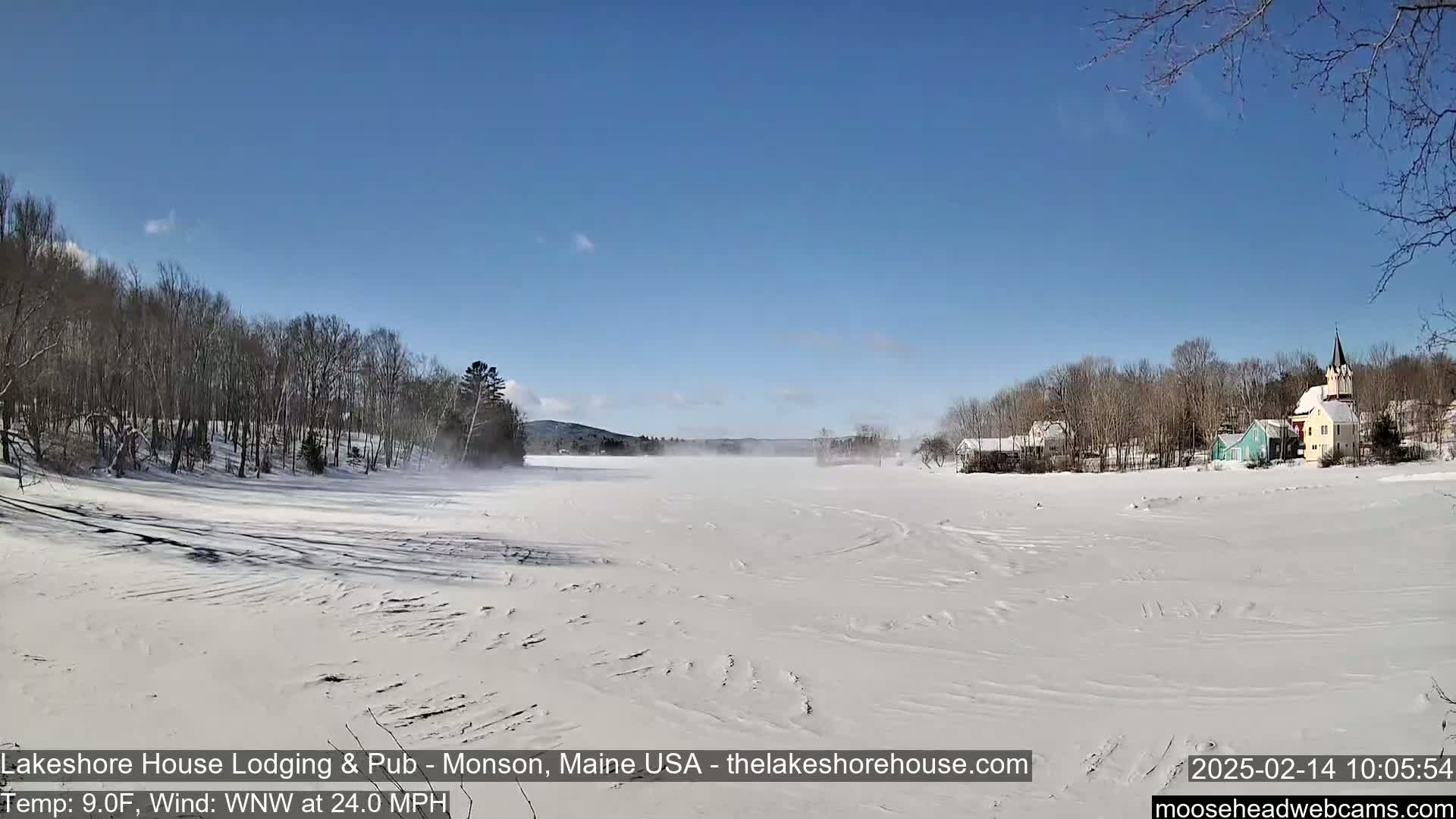 A snow-covered frozen lake stretches out before a line of snow-dusted trees and buildings under a clear blue sky with a strong wind.