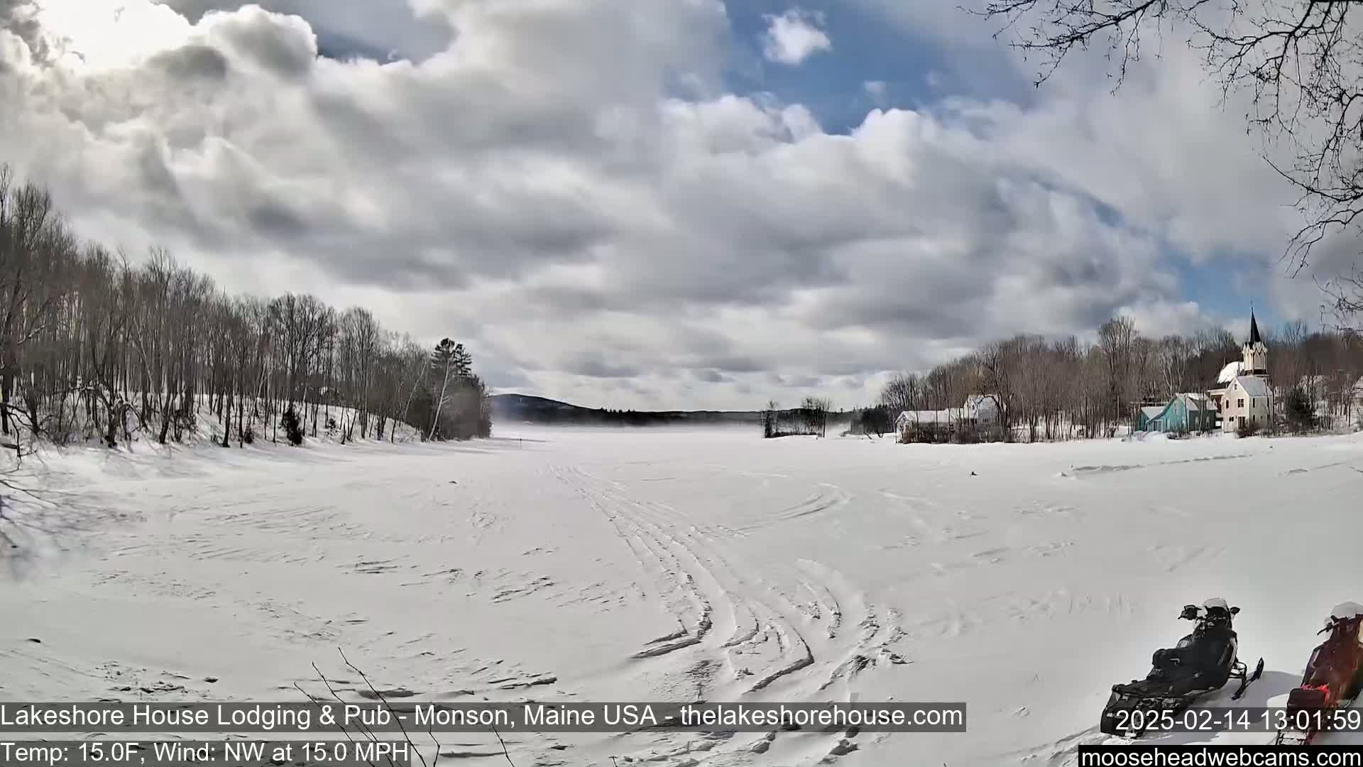 A snow-covered frozen lake with a village in the background under a cloudy sky, with a snowmobile parked on the ice and a northwest wind of 15 mph at a temperature of 15°F.