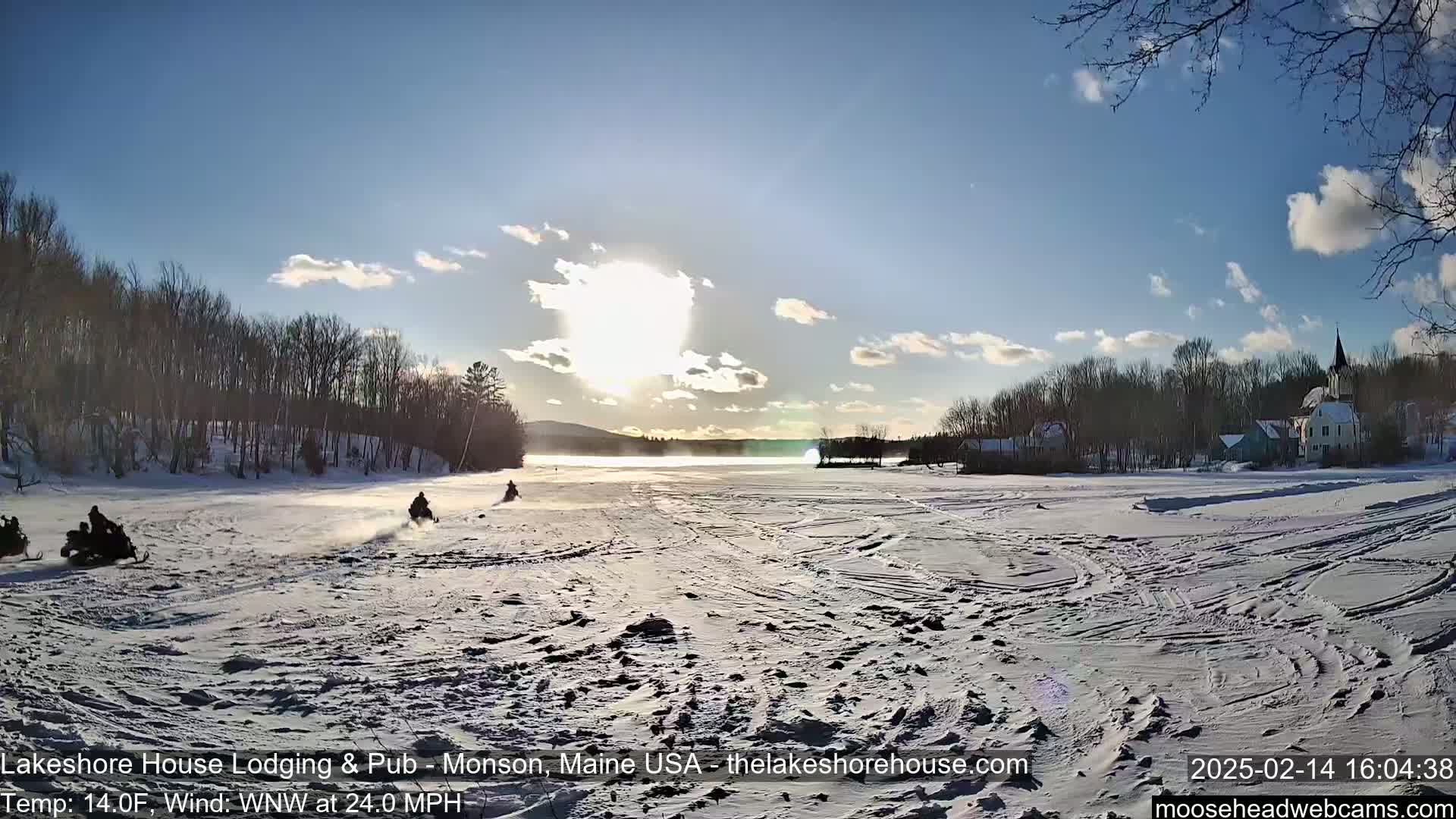 Snowmobiles travel across a snow-covered lake under a mostly sunny sky with some clouds, toward a village nestled among bare trees.