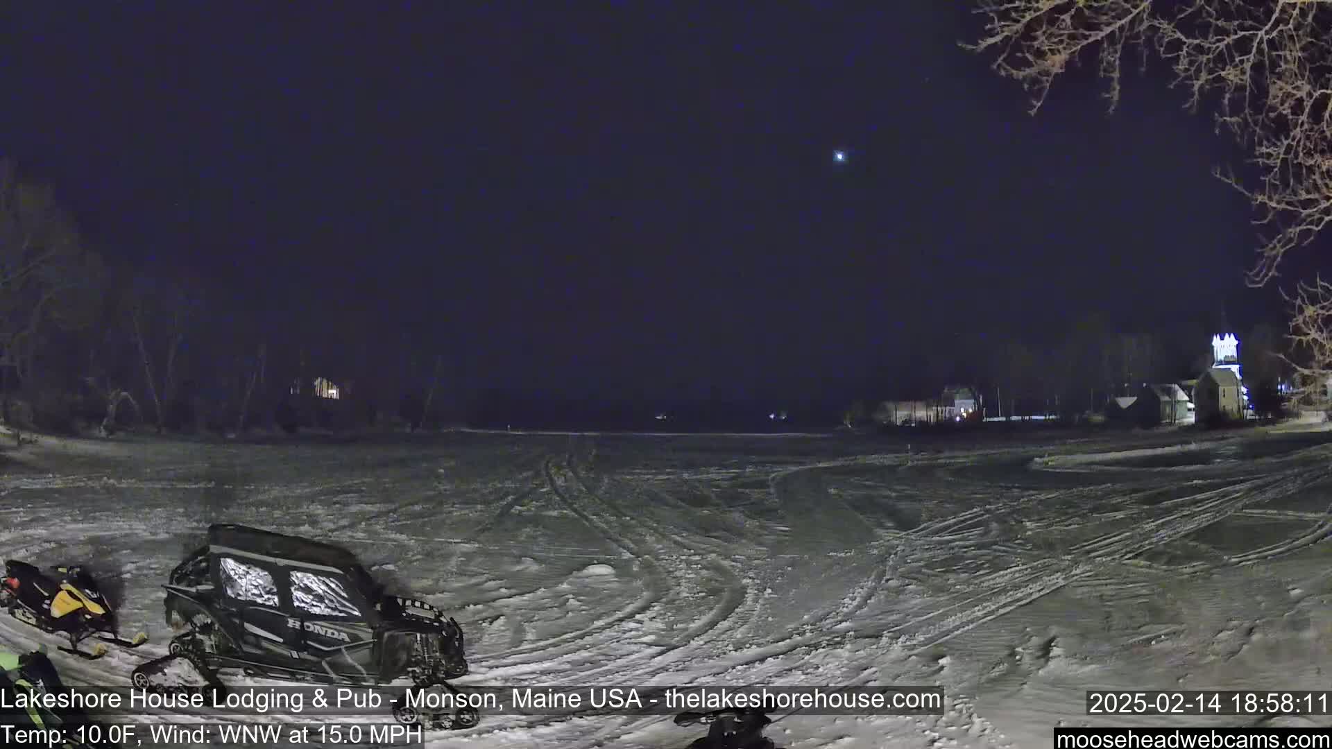 A snow-covered field at night, under a dark sky with a single bright star, features snowmobiles parked near a cluster of buildings in the distance.