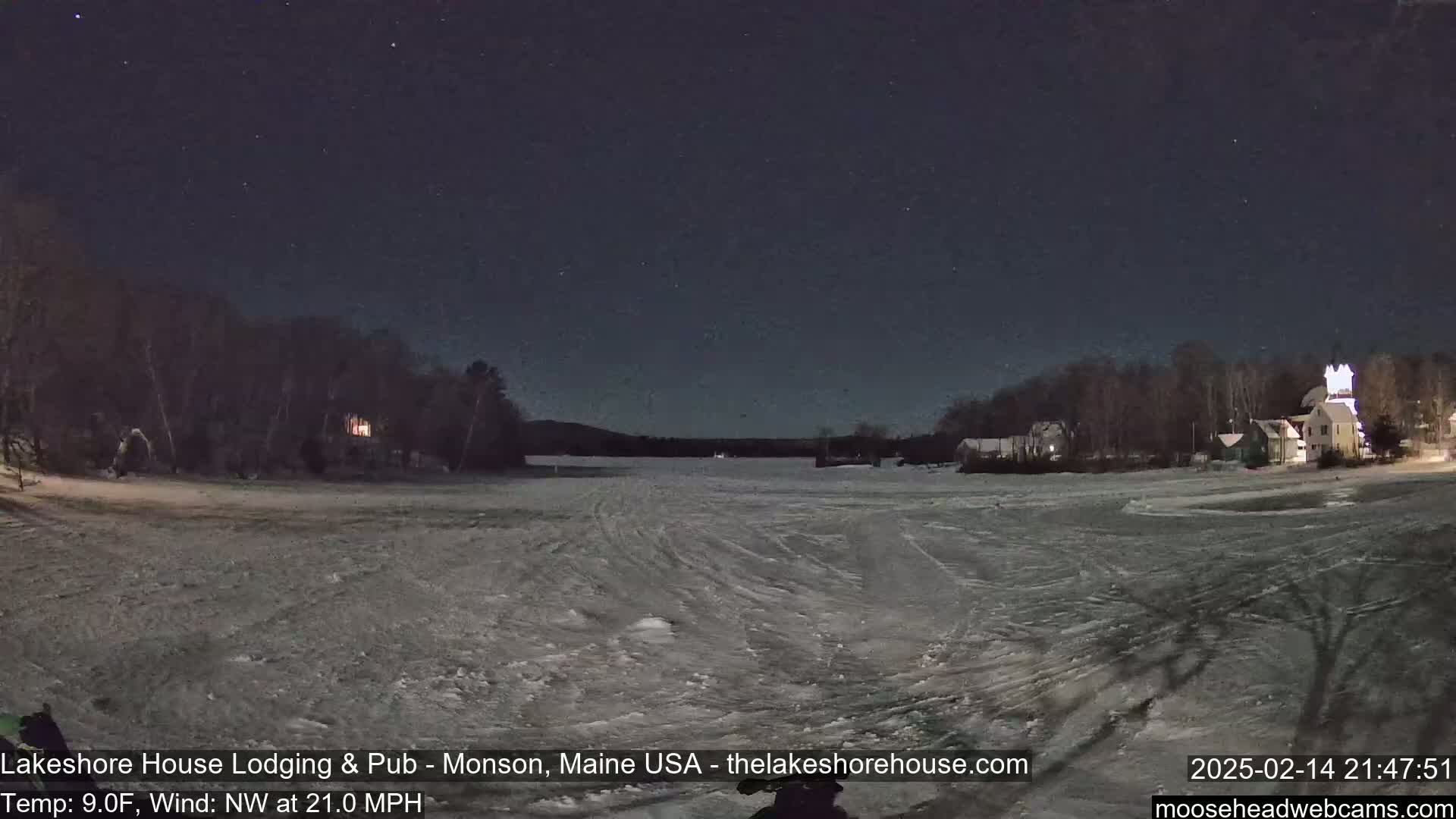 A frozen lake at night under a starry sky, with buildings and trees visible along the shoreline in the distance, and a northwest wind blowing at 21 miles per hour.