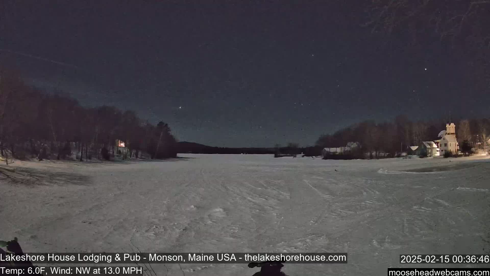 A snow-covered lake is seen at night under a clear, starry sky, with buildings and trees lining the shore.