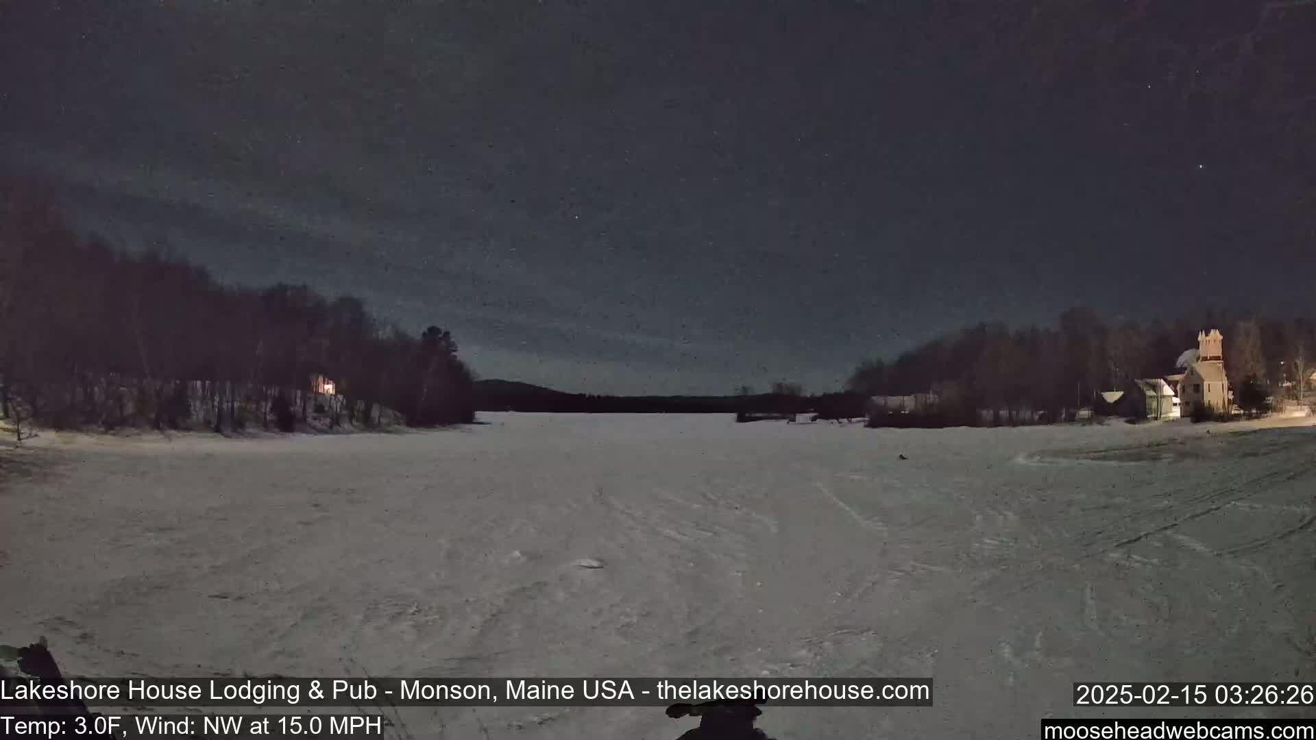 A snow-covered expanse stretches out under a night sky dotted with stars, with dark trees lining the sides and buildings visible in the distance under a 3°F temperature and 15 mph northwest wind.