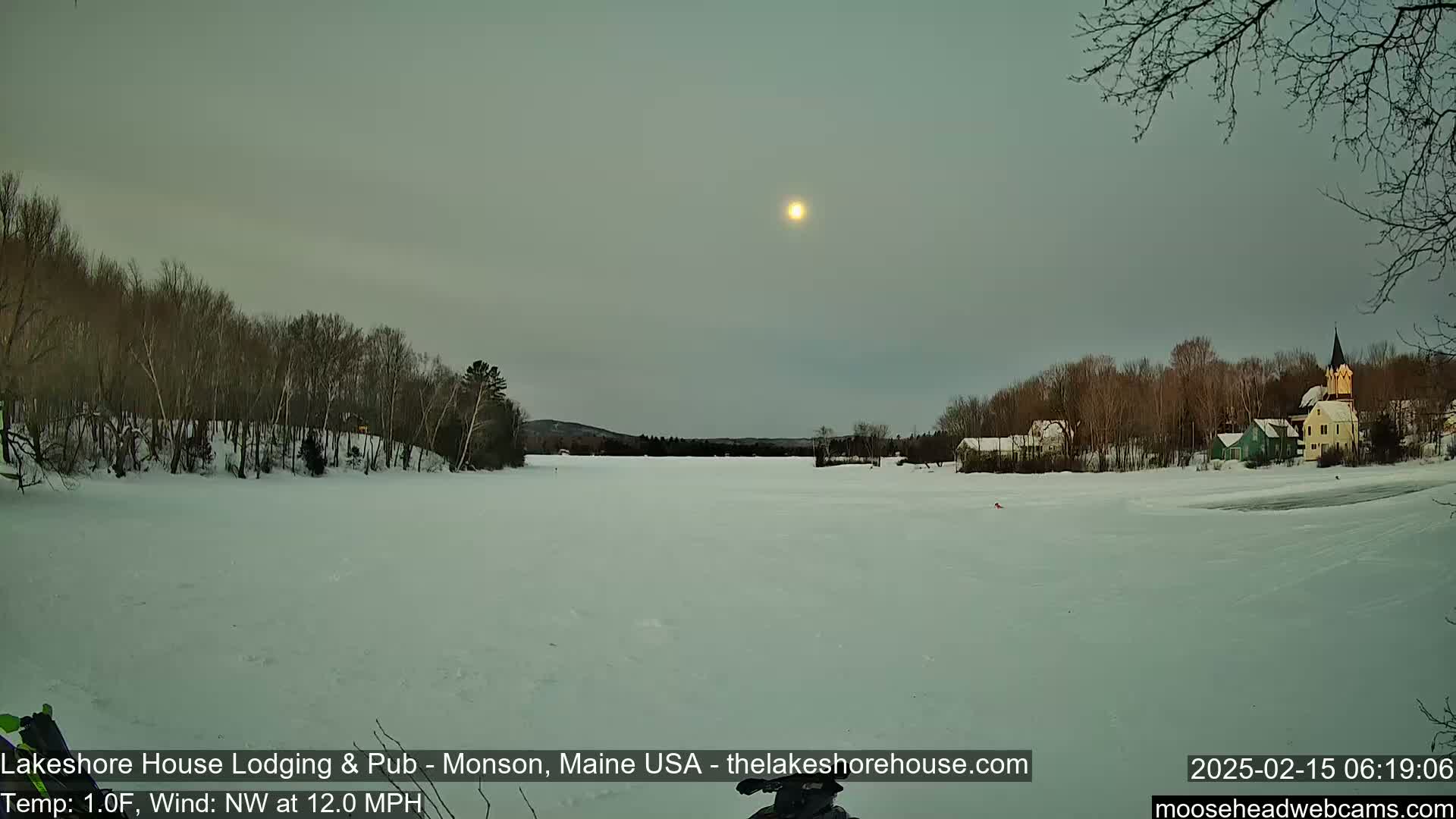 A snow-covered frozen lake stretches out before a small village under a pale, sunlit sky, with bare trees lining the shore.