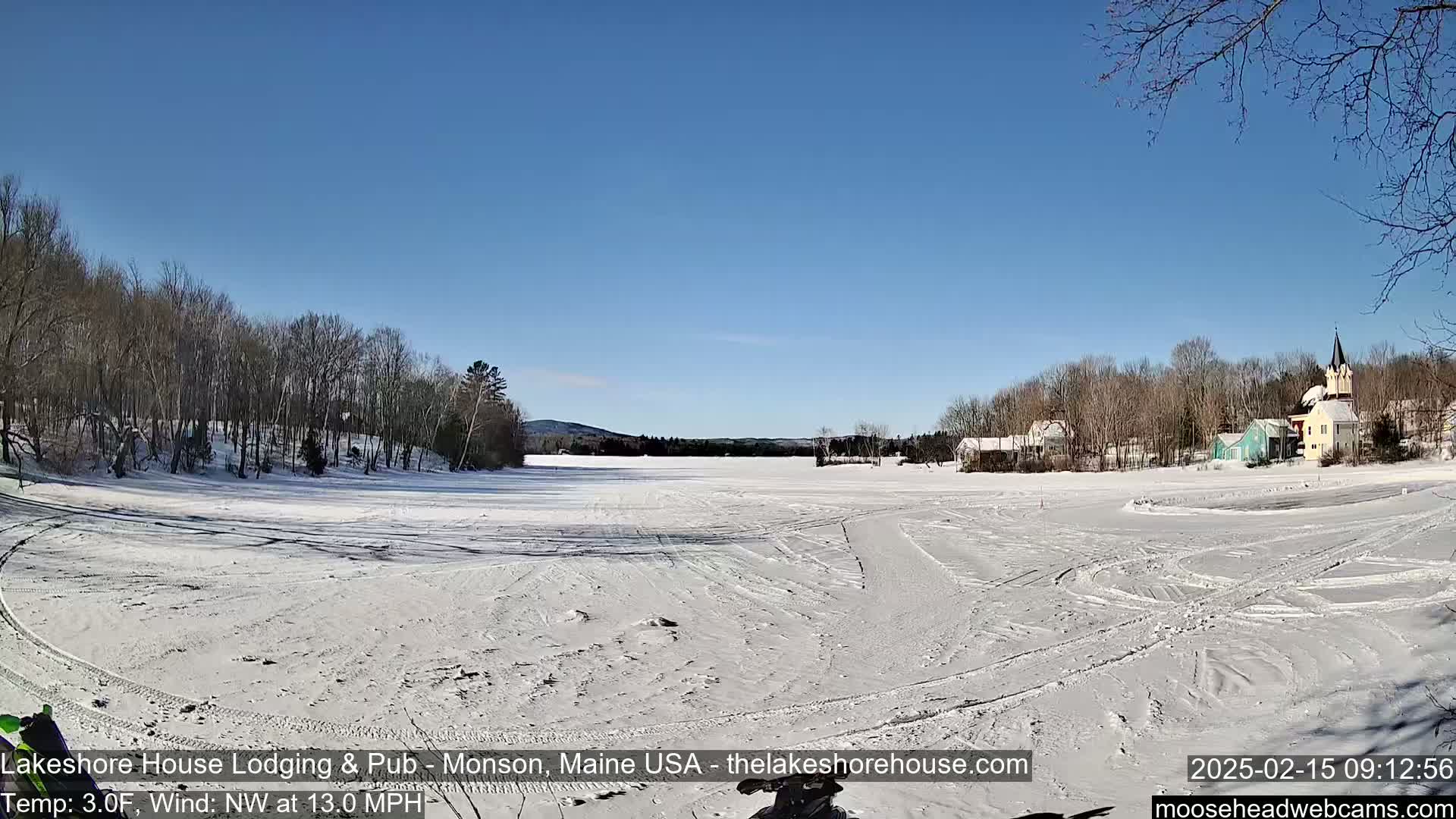 A snow-covered frozen lake stretches out to a small village under a clear blue sky, with a northwest wind blowing at 13 mph and a temperature of 3 degrees Fahrenheit.