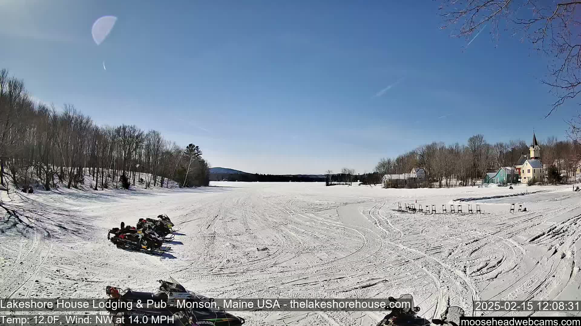 Several snowmobiles are parked on a snow-covered frozen lake under a sunny sky, with a village visible in the background.