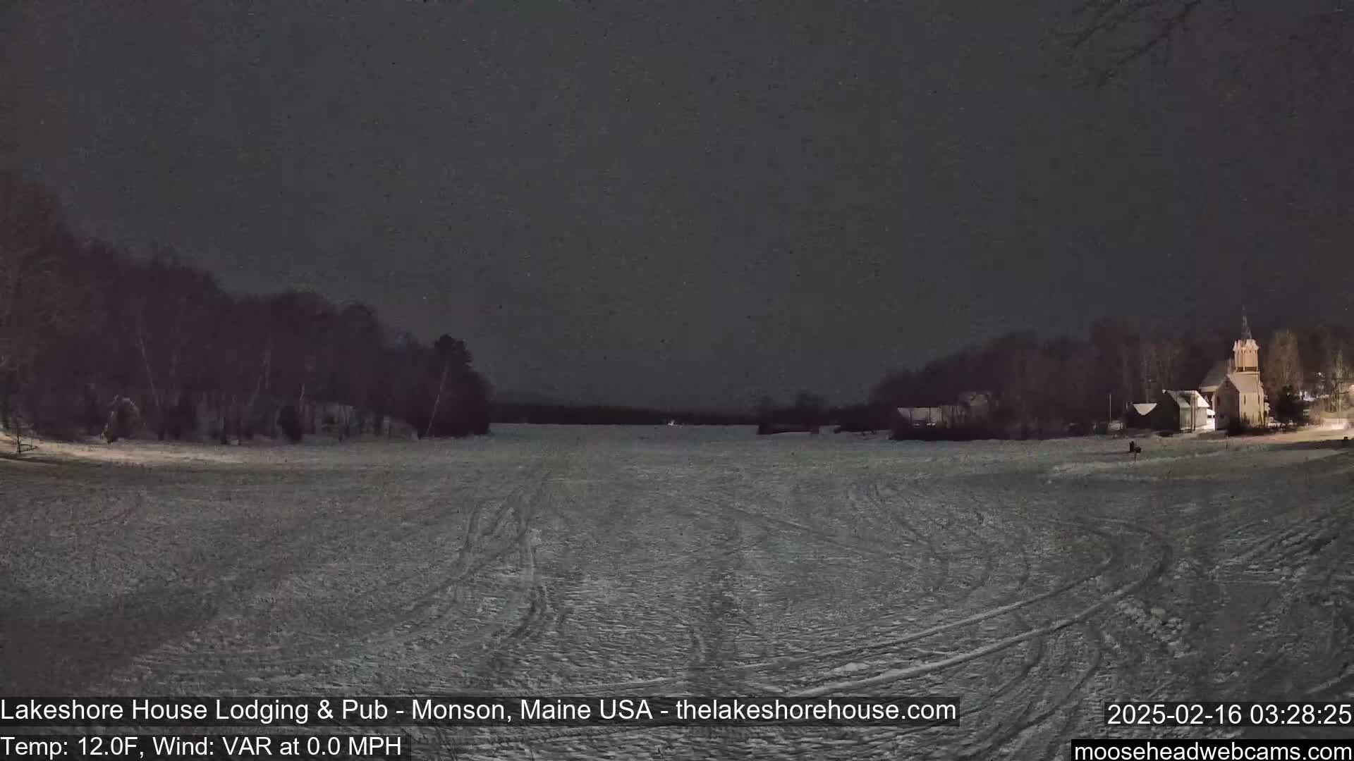 A snow-covered expanse stretches out under a dark night sky, with trees and buildings visible in the distance.
