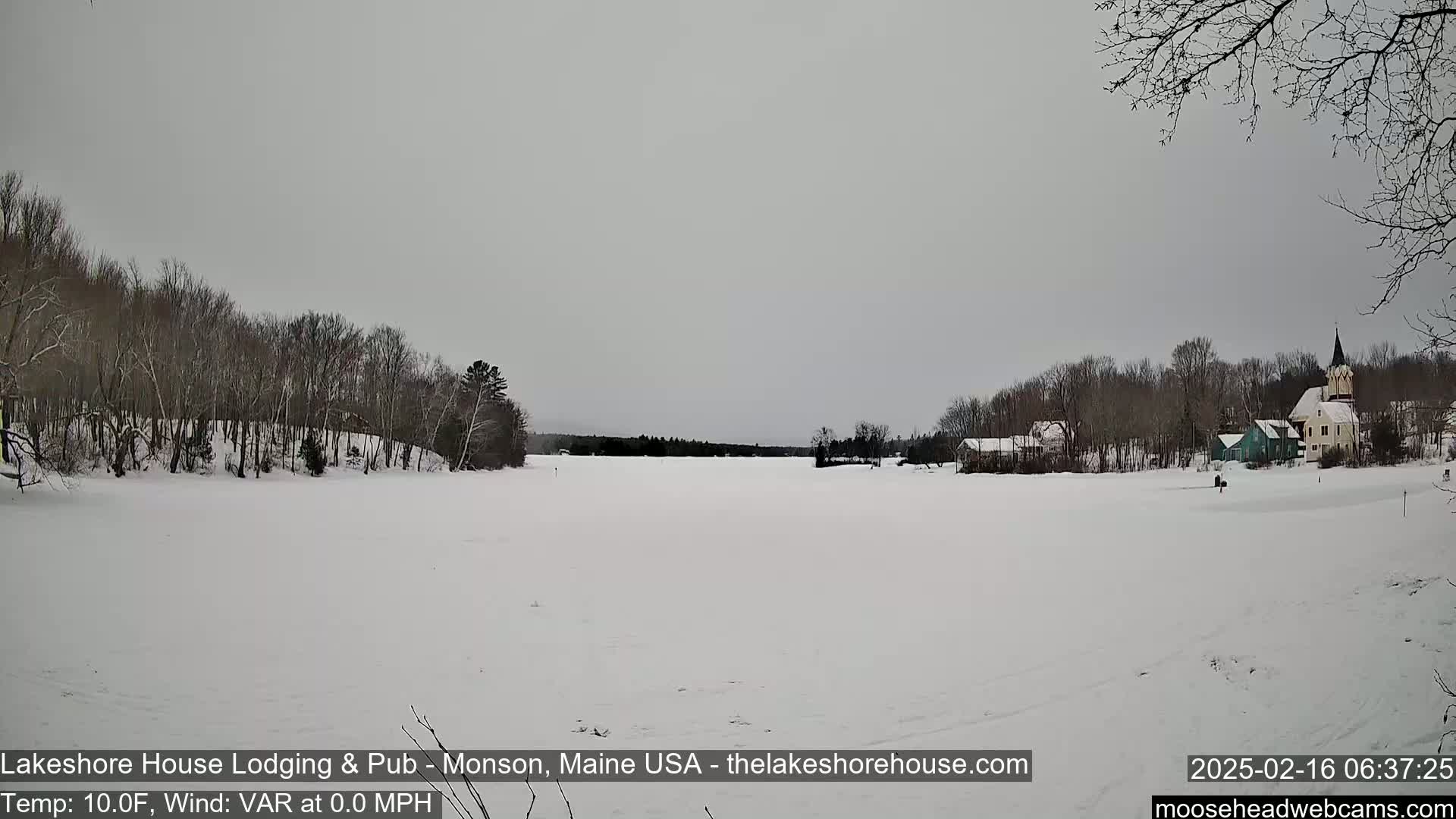 A snow-covered frozen lake stretches to a small village with buildings and a church steeple under an overcast sky.