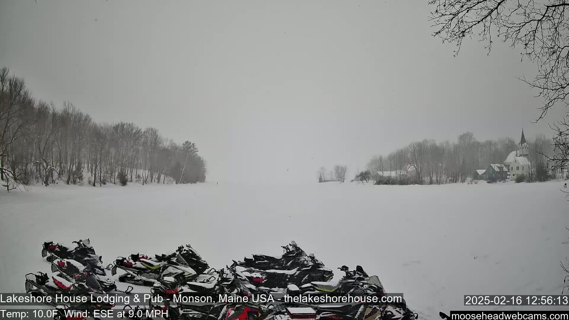A snowy scene shows a frozen lake with snowmobiles parked in the foreground and a village with a church steeple in the background, under a light snowfall.
