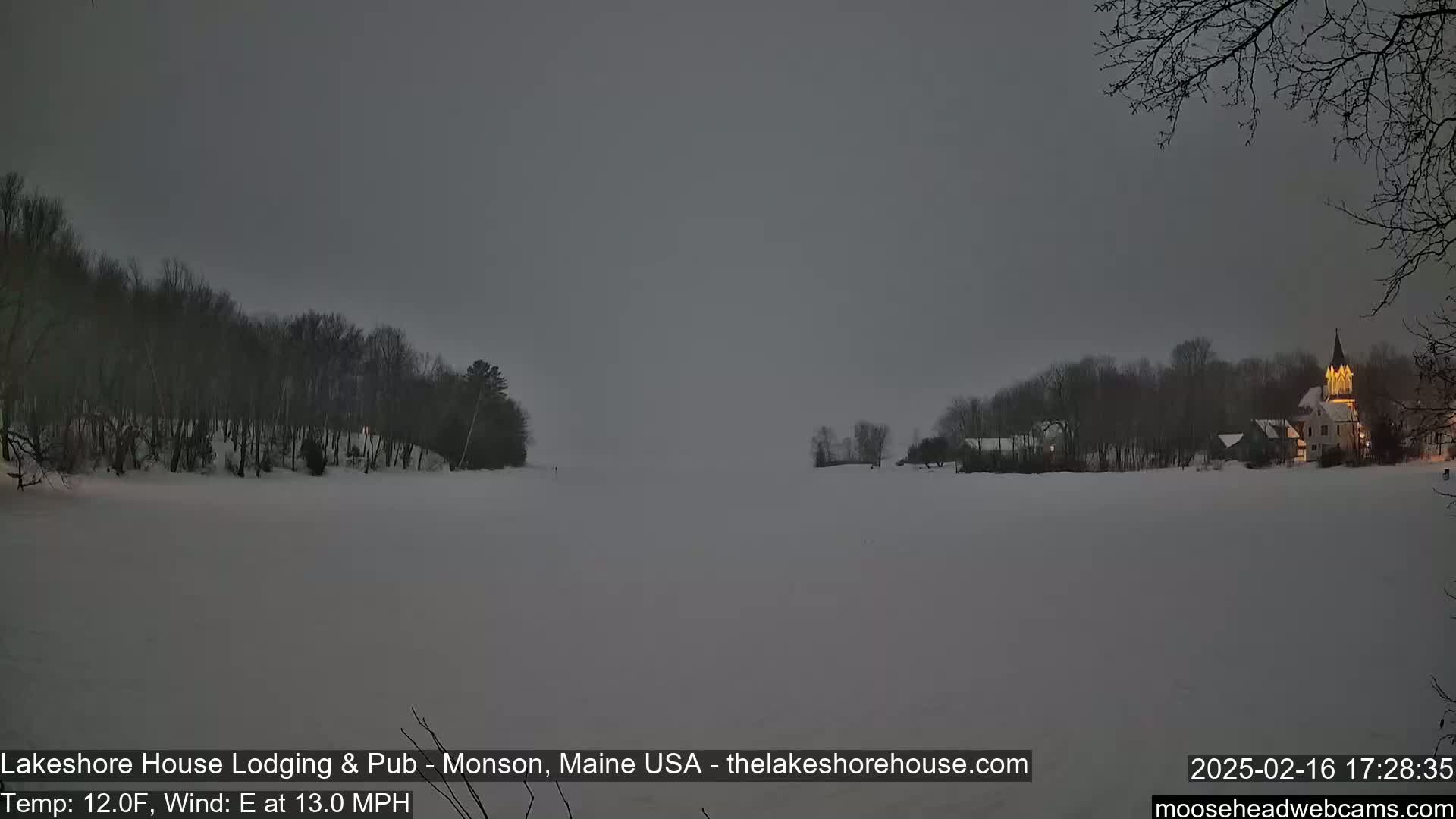 A snow-covered lake is seen at dusk, with dark, snow-covered trees lining the shore and a lit building visible in the distance under a gray sky.