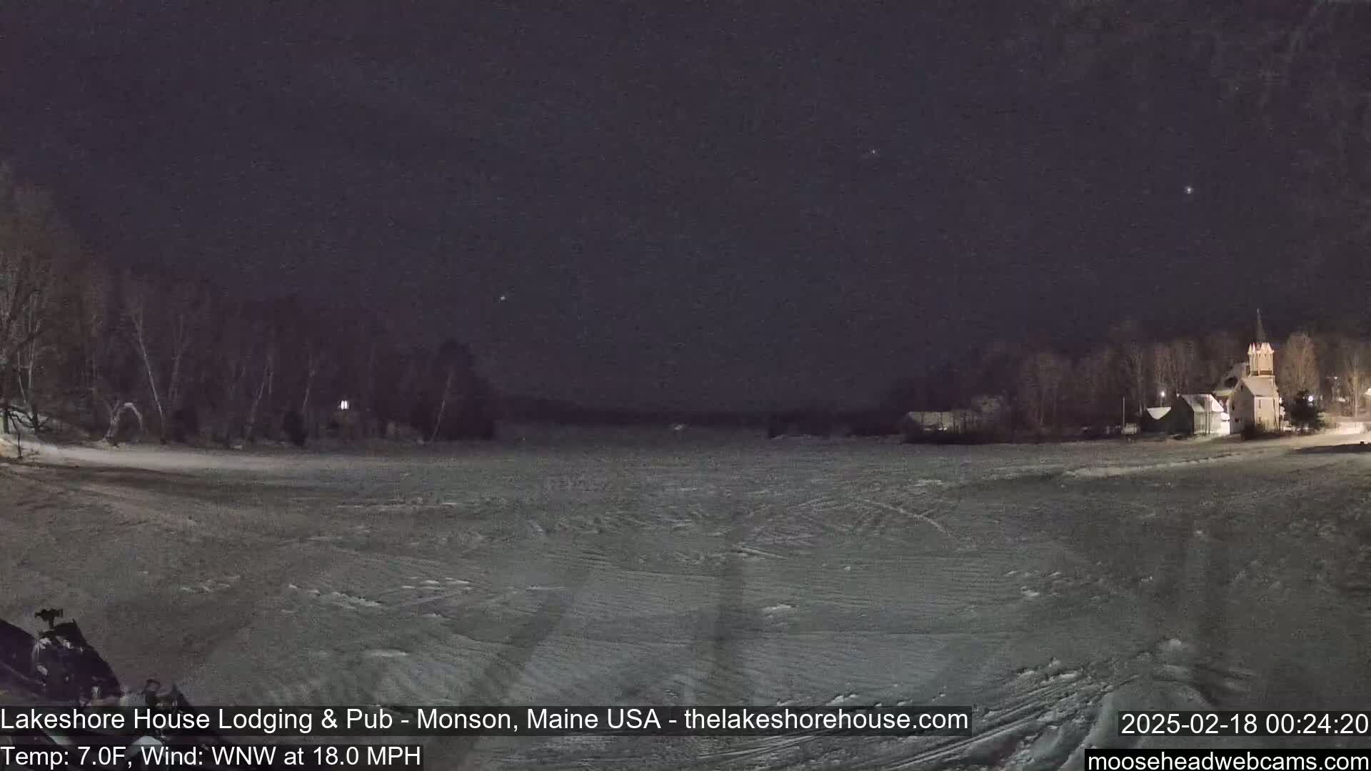 A snow-covered expanse at night, illuminated by faint lights from buildings in the distance, under a dark sky with a few visible stars, and a wind of 18 mph.