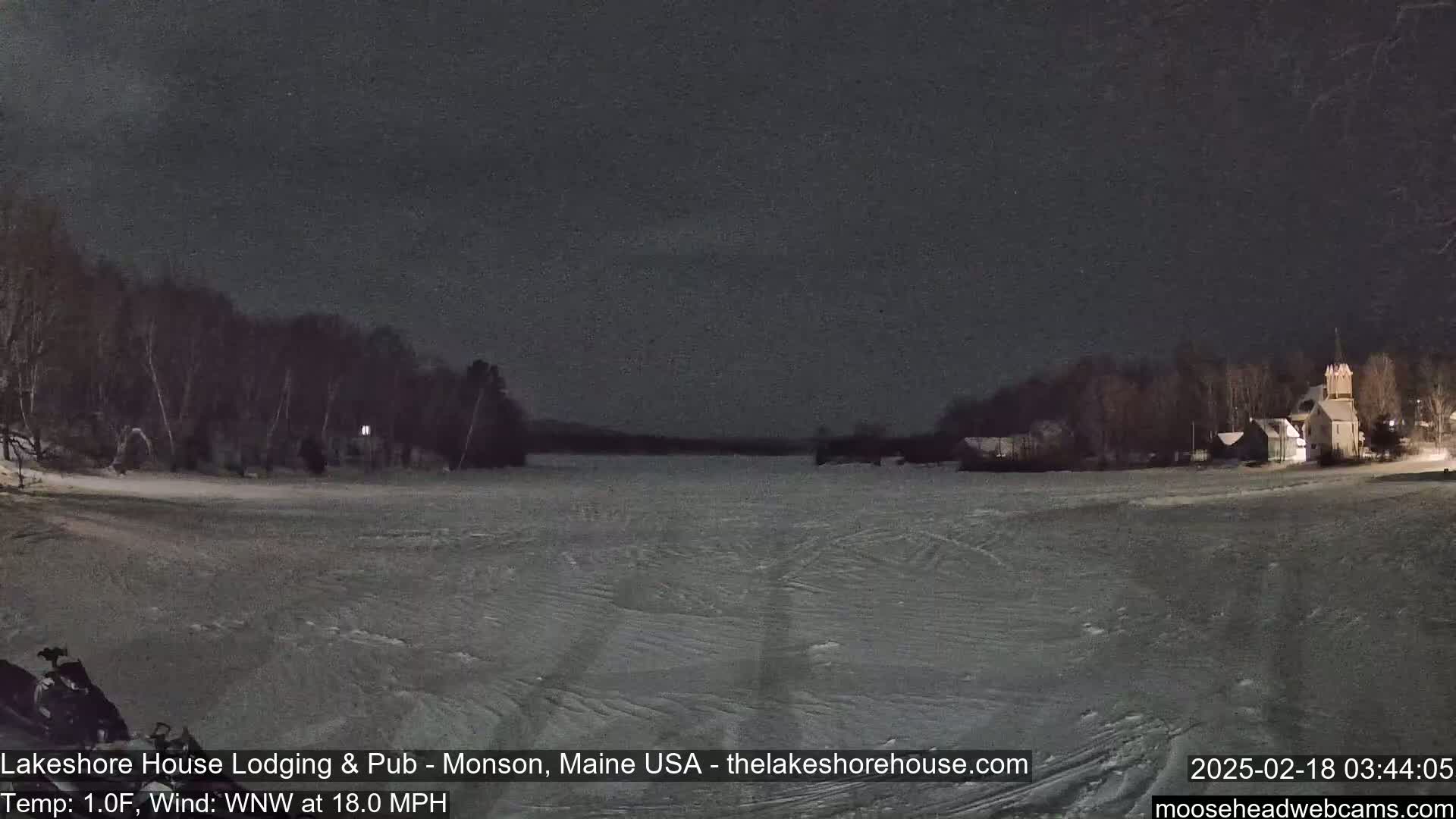 A snow-covered frozen lake is seen at night under a dark sky, with a sparsely lit shoreline featuring trees and buildings in the distance.