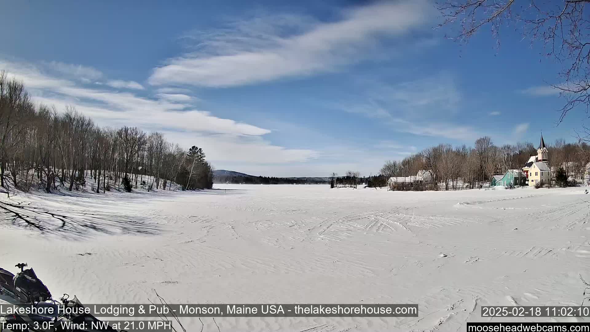 A snow-covered frozen lake stretches to a village of houses and a church under a mostly sunny sky with some scattered clouds.