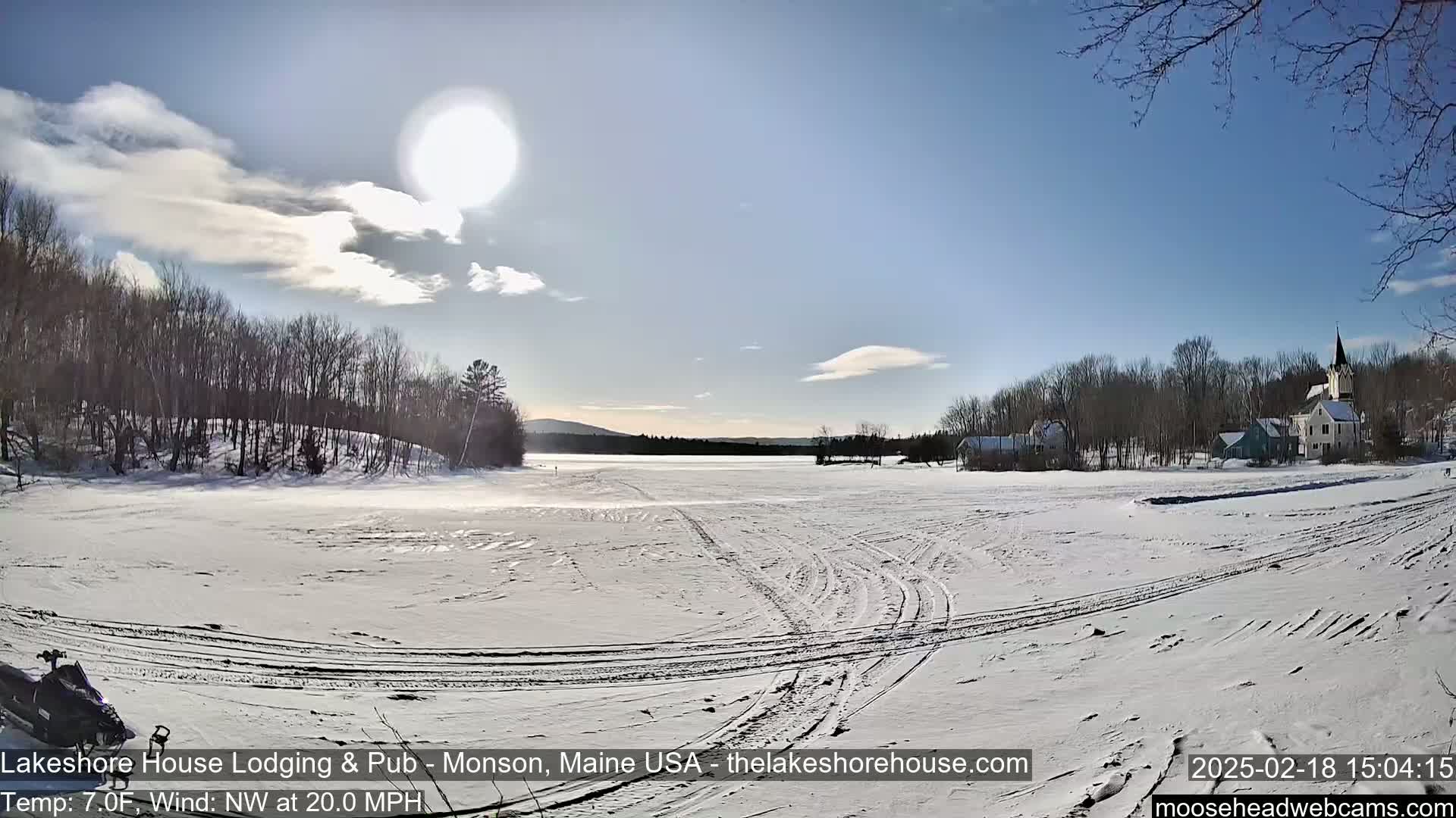 A snow-covered frozen lake with tire tracks, surrounded by bare trees and a small village under a bright, sunny sky with a few clouds.