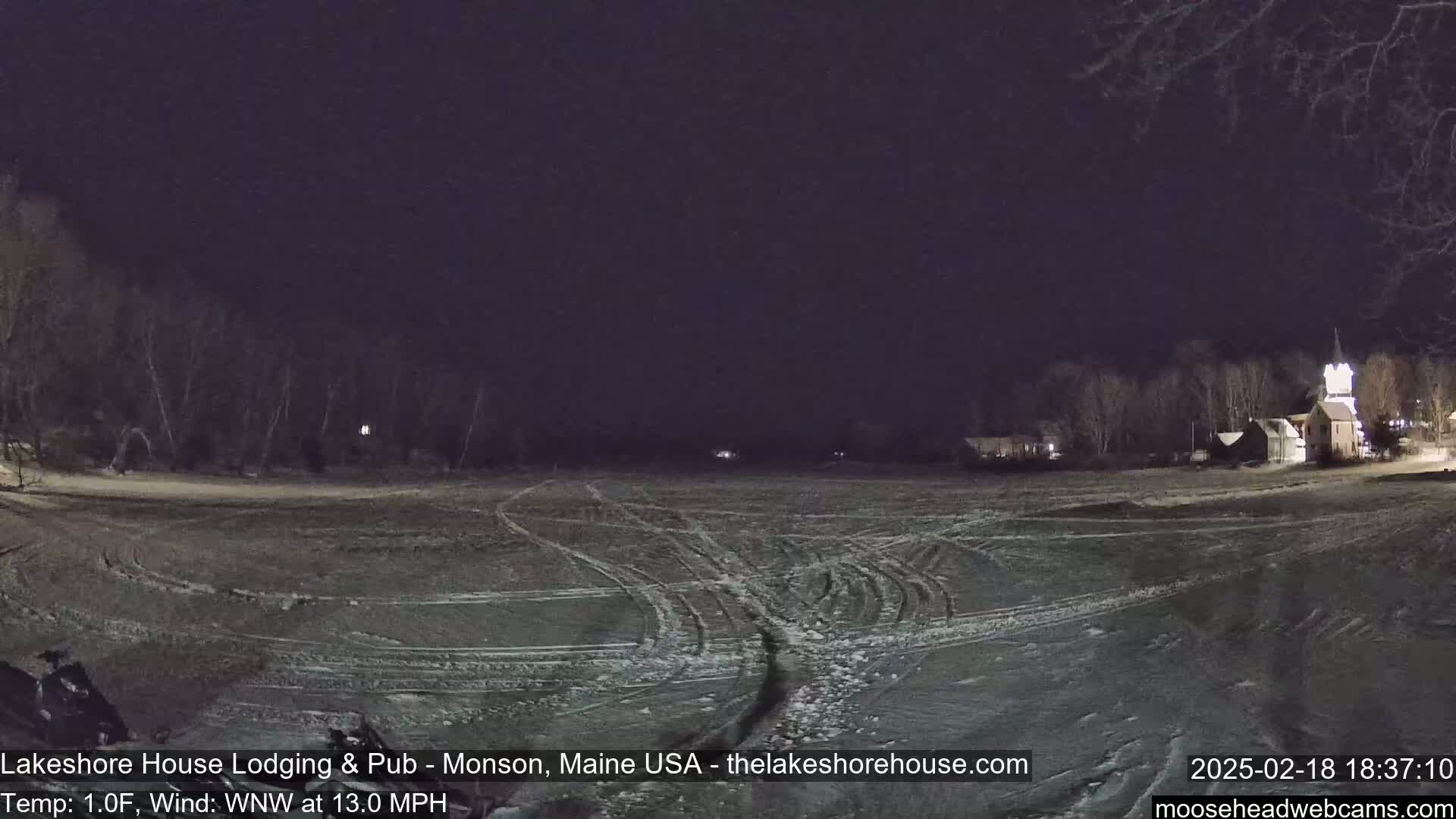 A snow-covered field at night, with tire tracks visible and a building illuminated in the background under a dark sky.