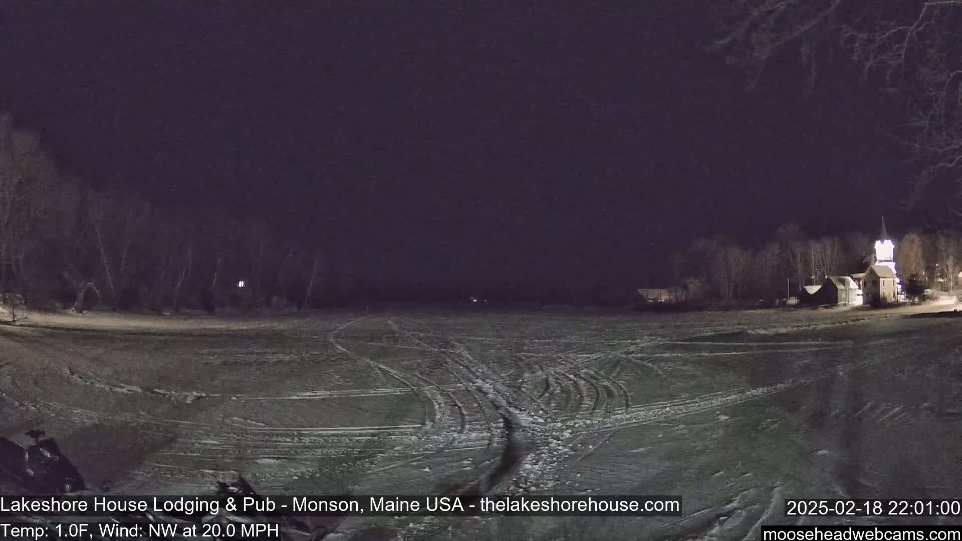 A snow-covered, flat area with tire tracks is visible at night, under a dark sky, with a building and trees in the background.
