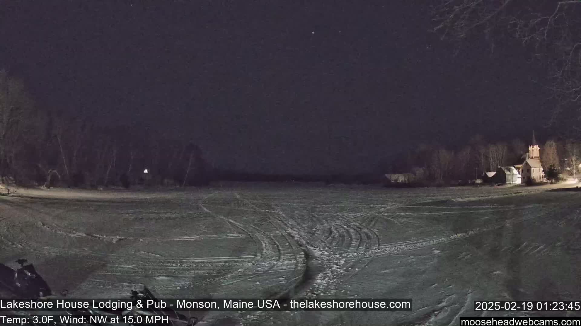 A snow-covered field at night, under a dark sky with a few stars, shows tire tracks and buildings in the distance.