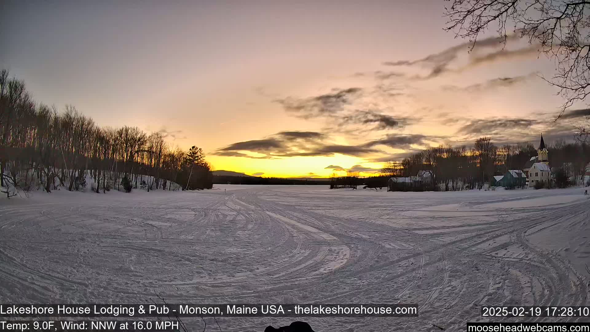 A frozen lake, with tire tracks crisscrossing its surface, shows a sunset over a snow-covered landscape featuring trees and buildings, under a partly cloudy sky with a 9°F temperature and a 16 mph northwest wind.