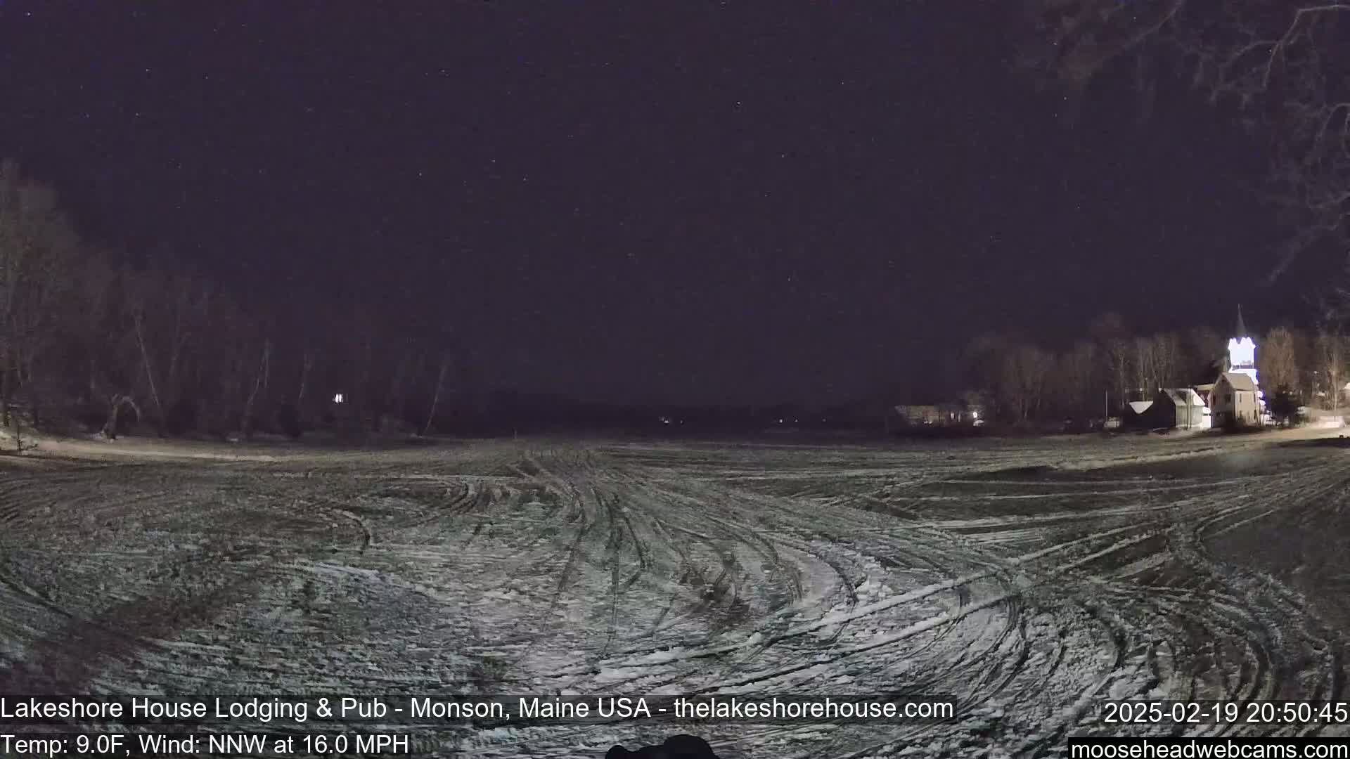 A snow-covered field with tire tracks under a dark, starry night sky, with a faint light from buildings in the distance.