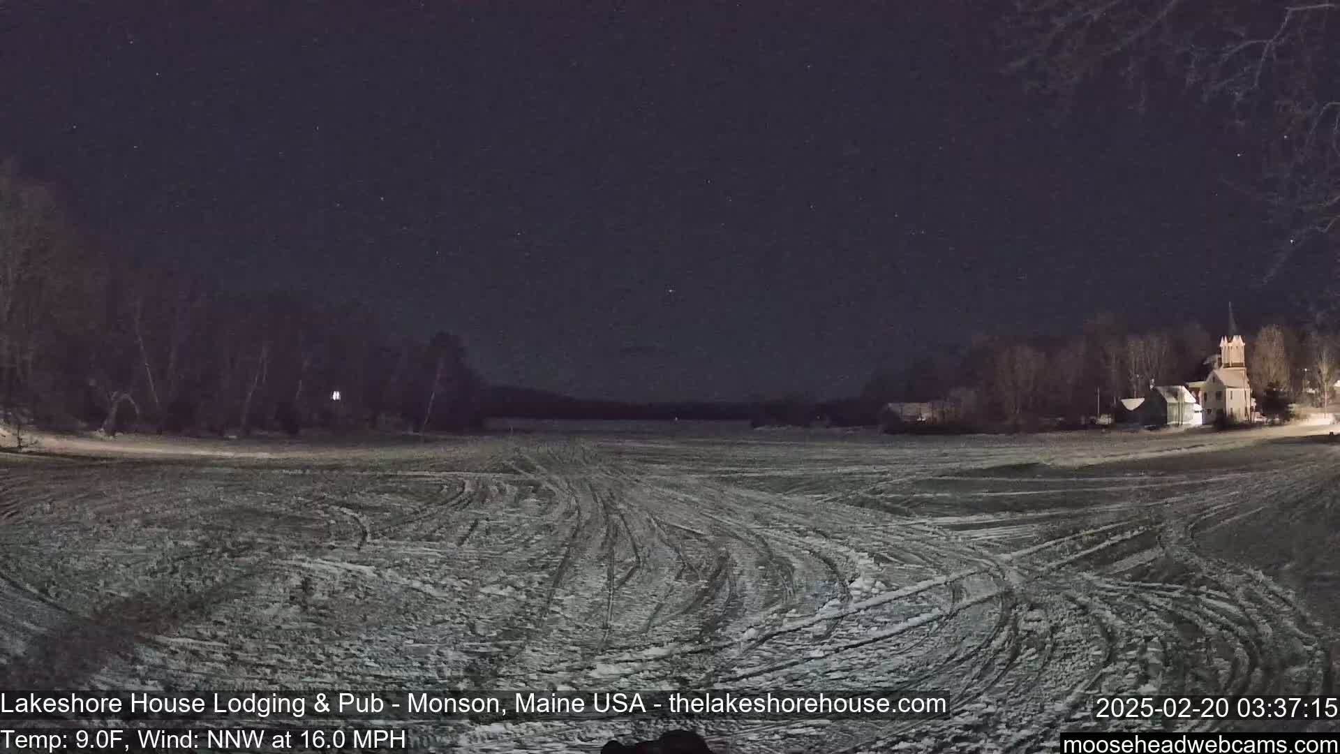 A snow-covered, vehicle-tracked field at night under a starry sky shows distant buildings and trees.
