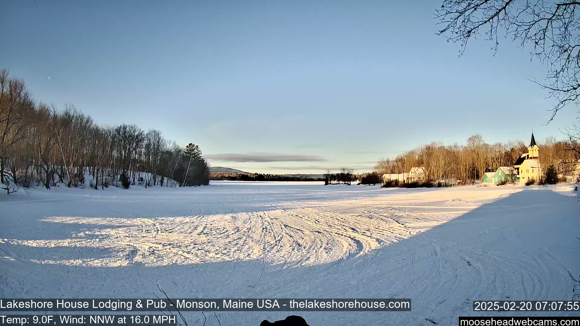 A frozen lake, covered in tire tracks, stretches toward a village of buildings under a clear, sunny sky; bare trees line the lake's edge.