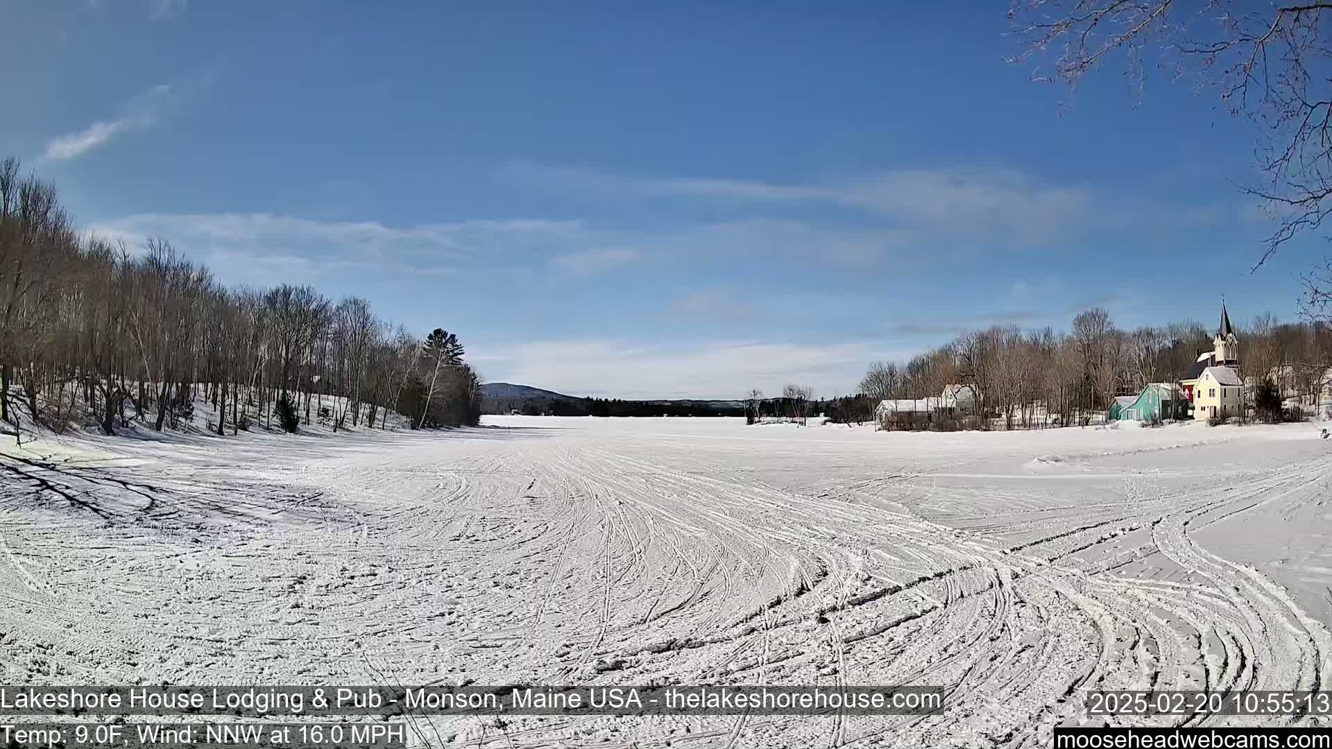 A snow-covered frozen lake with tracks from vehicles or snowmobiles, under a mostly clear blue sky, shows a small village in the distance.