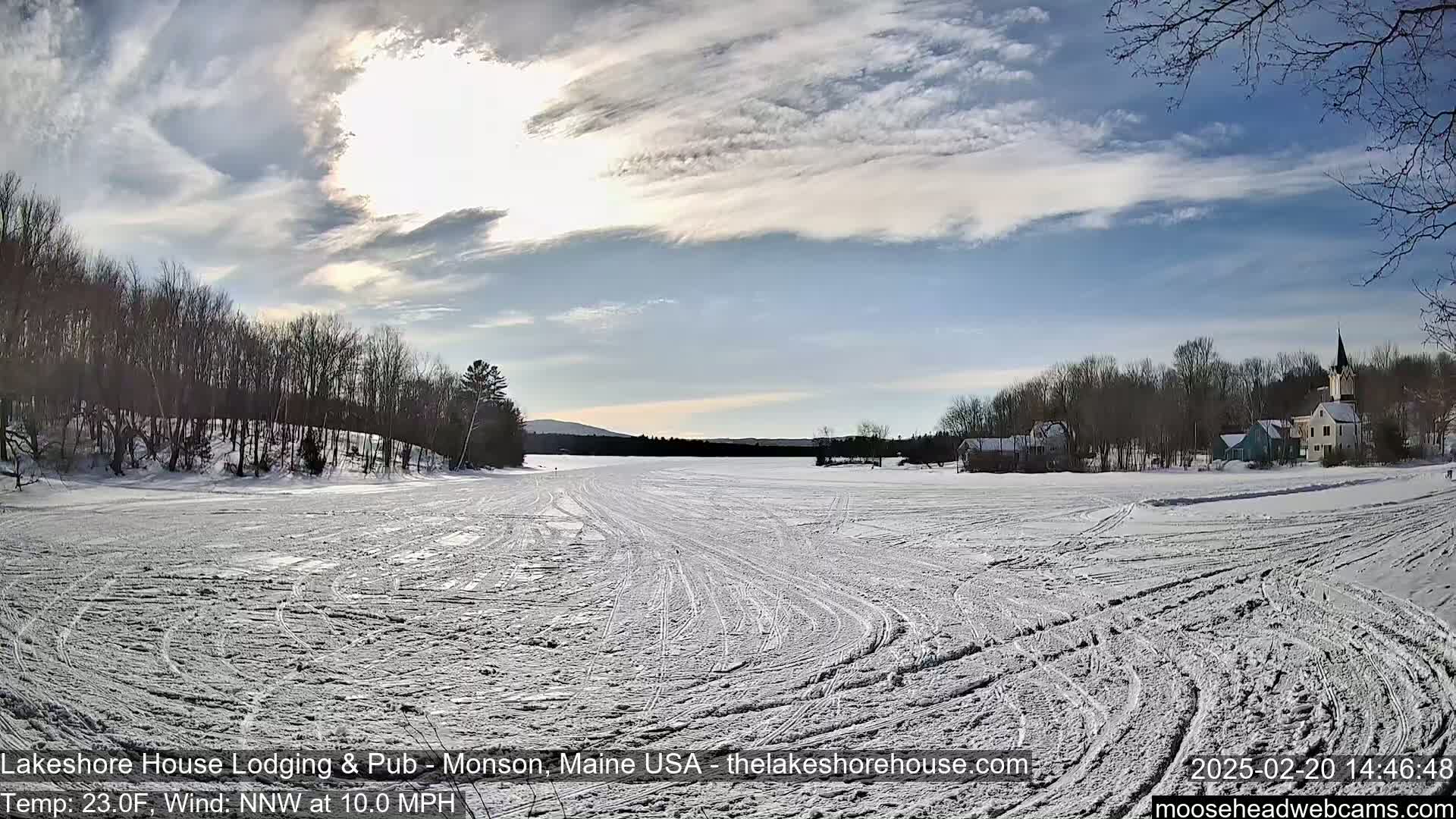 A snow-covered frozen lake with tire tracks, under a partly sunny sky, shows a village with buildings and trees in the distance.