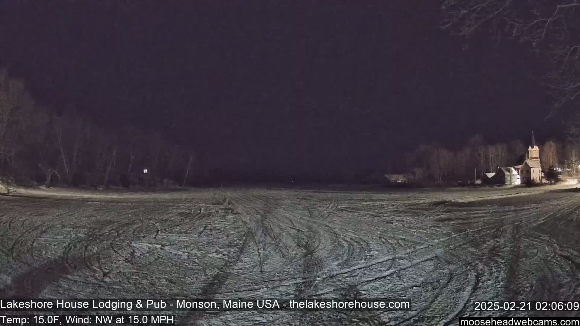 A snow-covered field with tire tracks under a dark night sky shows a building and trees in the distance, with a northwest wind at 15 mph and a temperature of 15 degrees Fahrenheit.