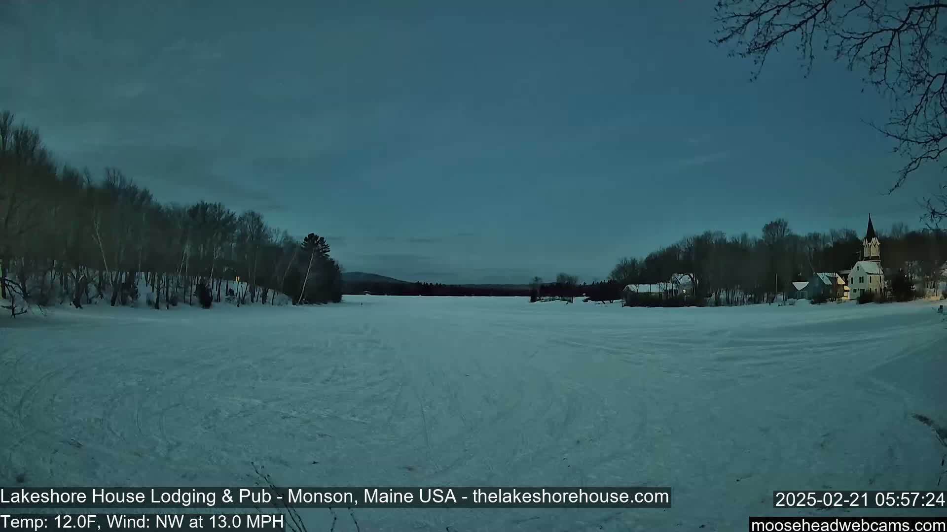 A snow-covered frozen lake stretches to a treeline and buildings under a dark, twilight sky with a northwest wind.