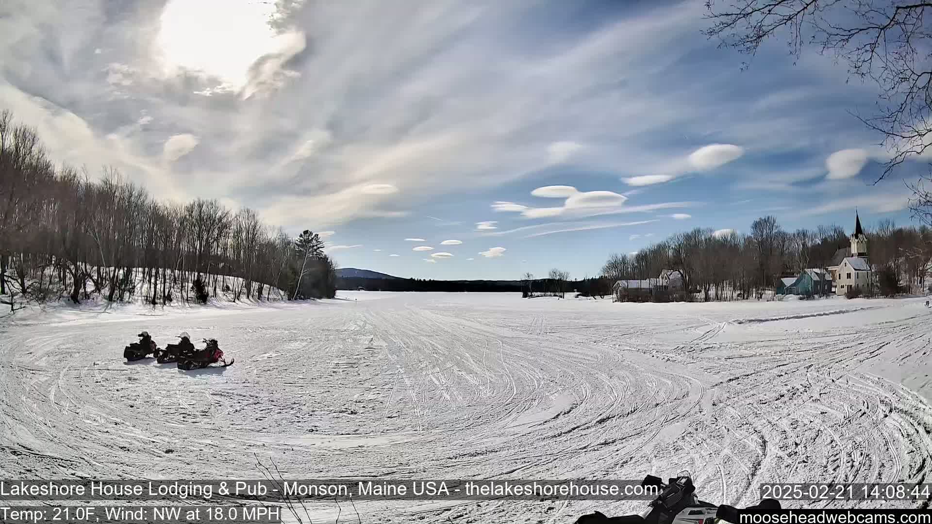 Three snowmobiles are parked on a snow-covered frozen lake under a partly sunny sky with lenticular clouds, and a small village is visible in the background.