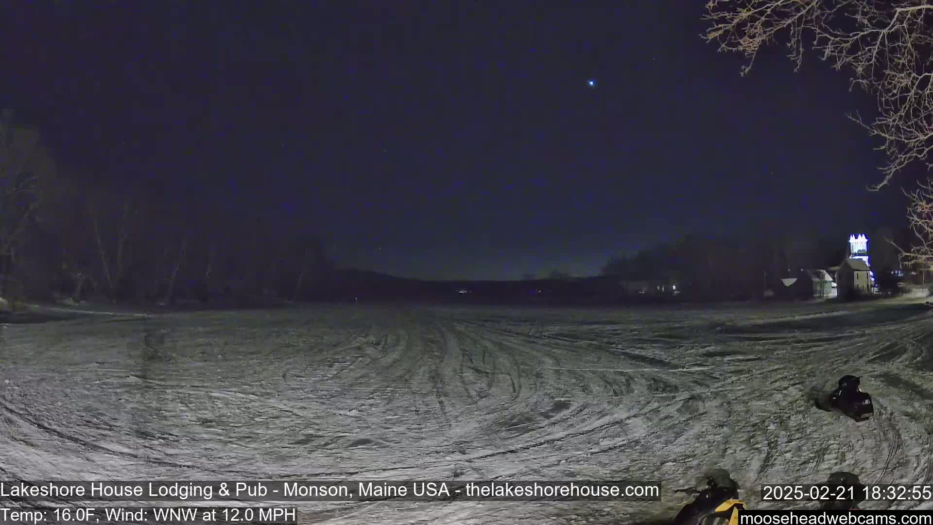 A snow-covered field with tire tracks under a dark, starry night sky, with a few buildings visible in the distance and a snowmobile in the lower right corner, with a wind of 12 mph.