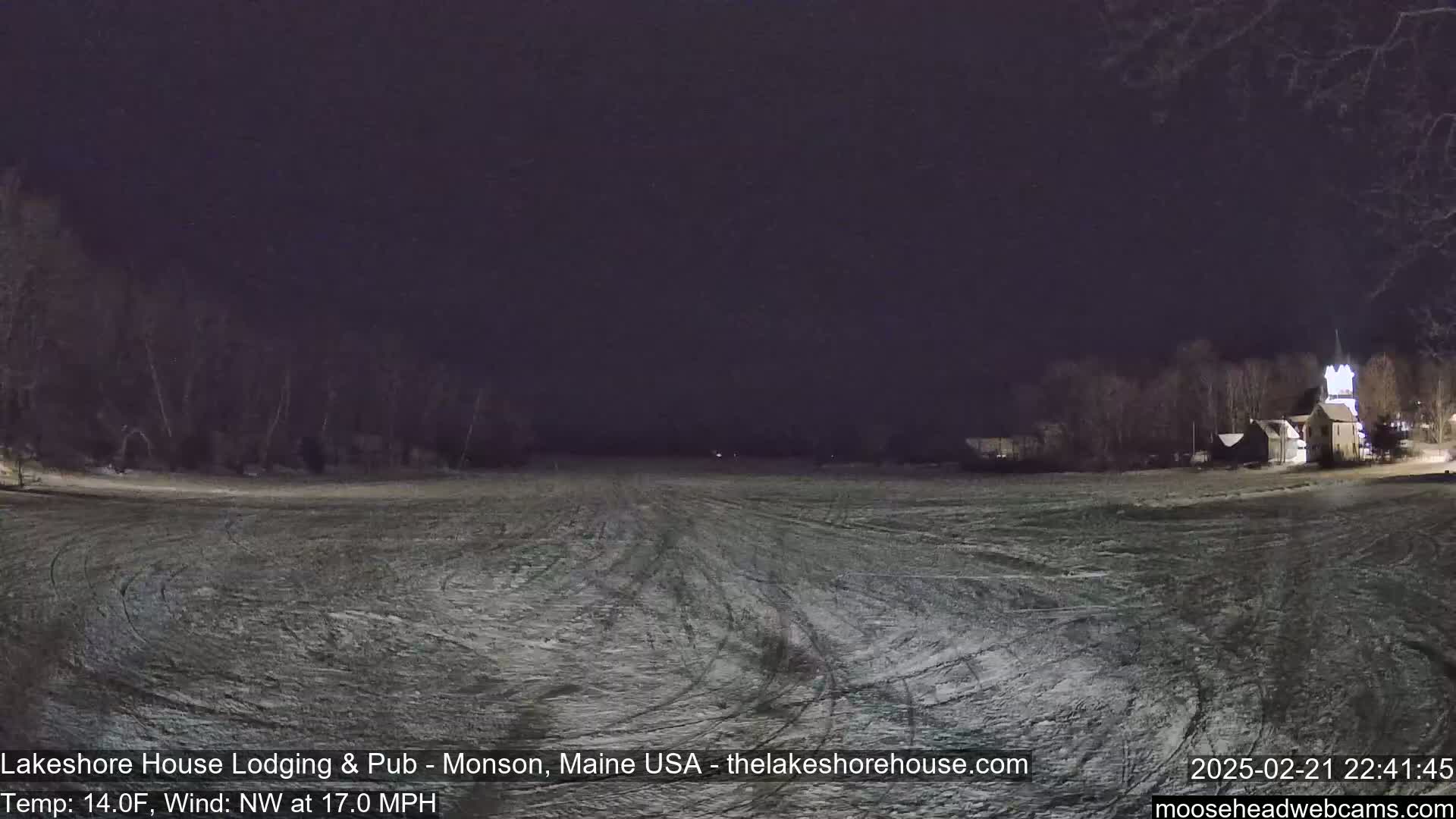 A snow-covered field at night with a building visible in the distance under a dark sky, with a northwest wind at 17 mph and a temperature of 14°F.