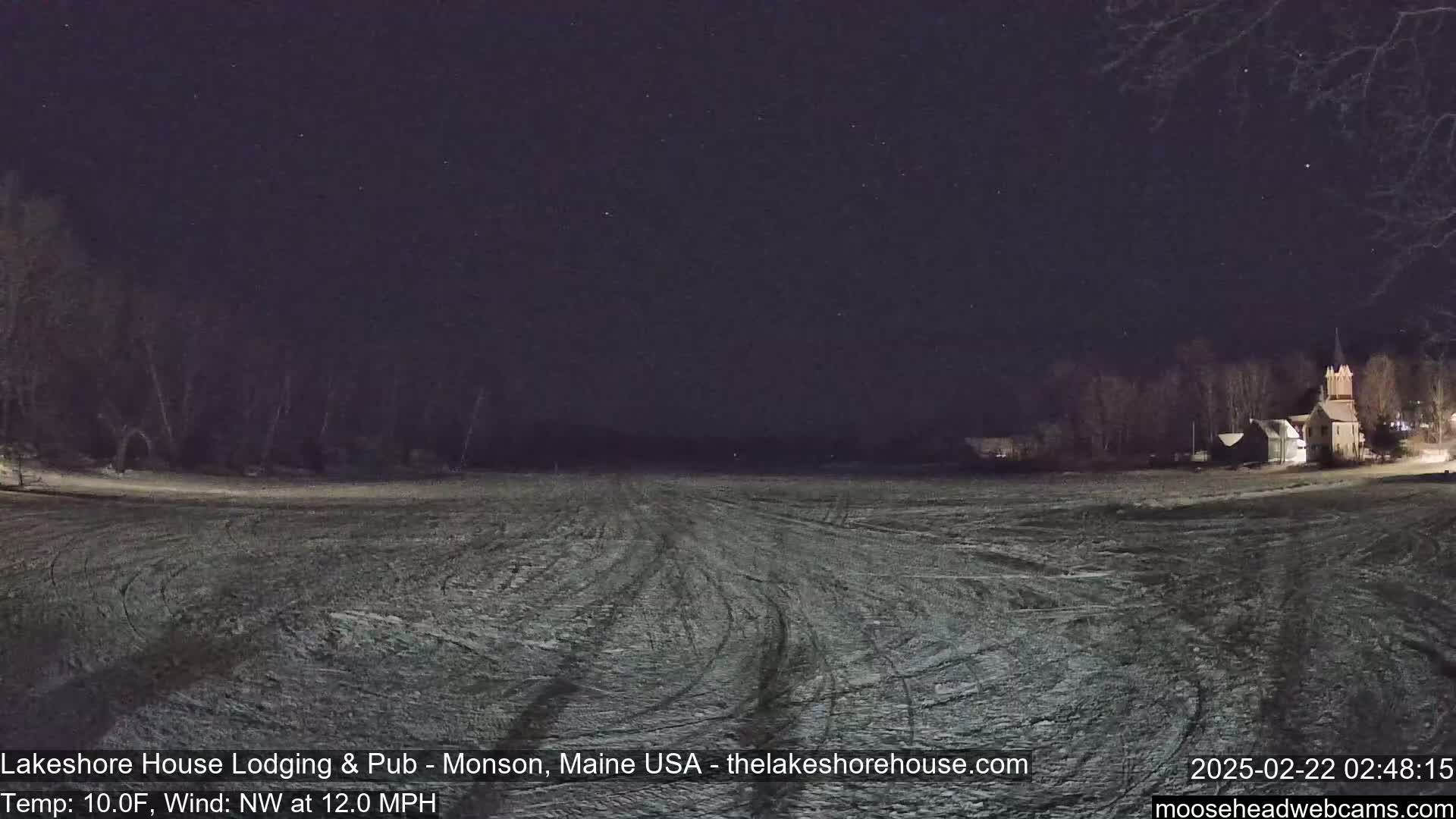 A snow-covered field with tire tracks under a starry night sky, with a building visible in the distance.