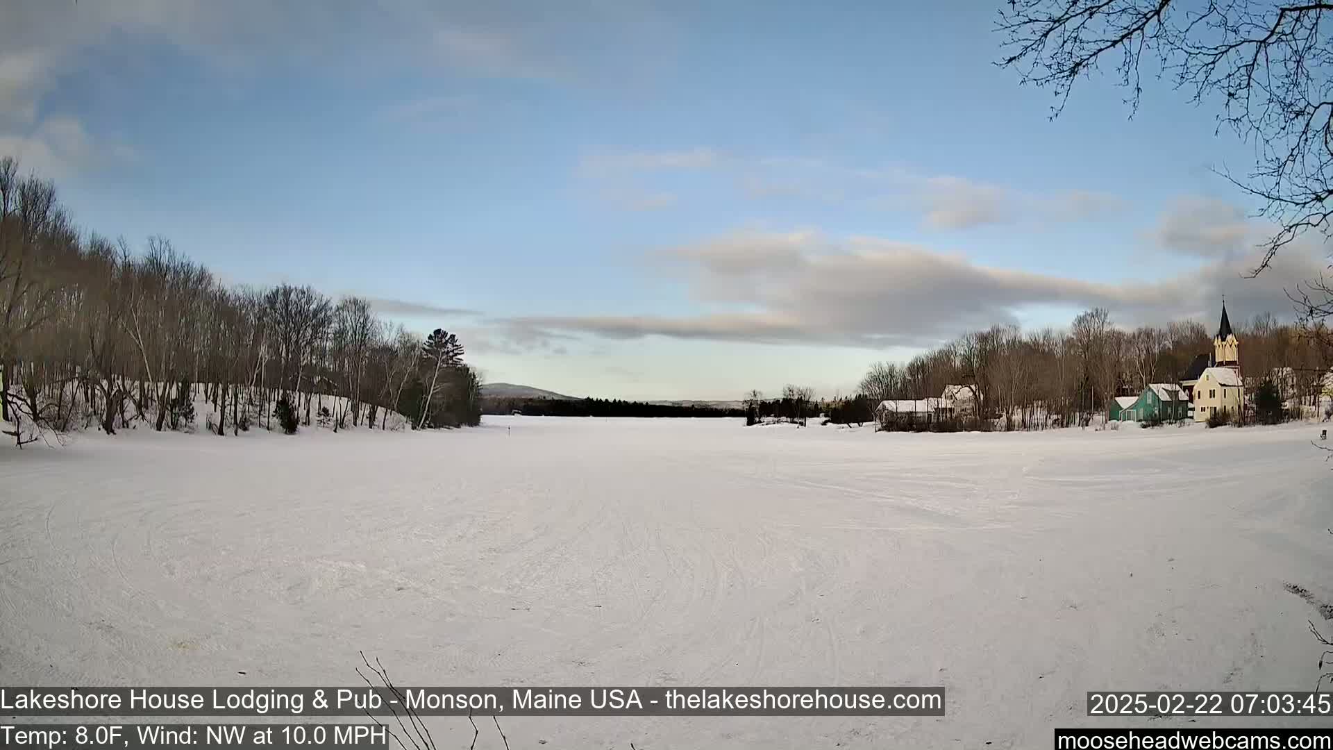 A snow-covered frozen lake stretches to a village of houses and a church under a mostly clear, light blue sky.