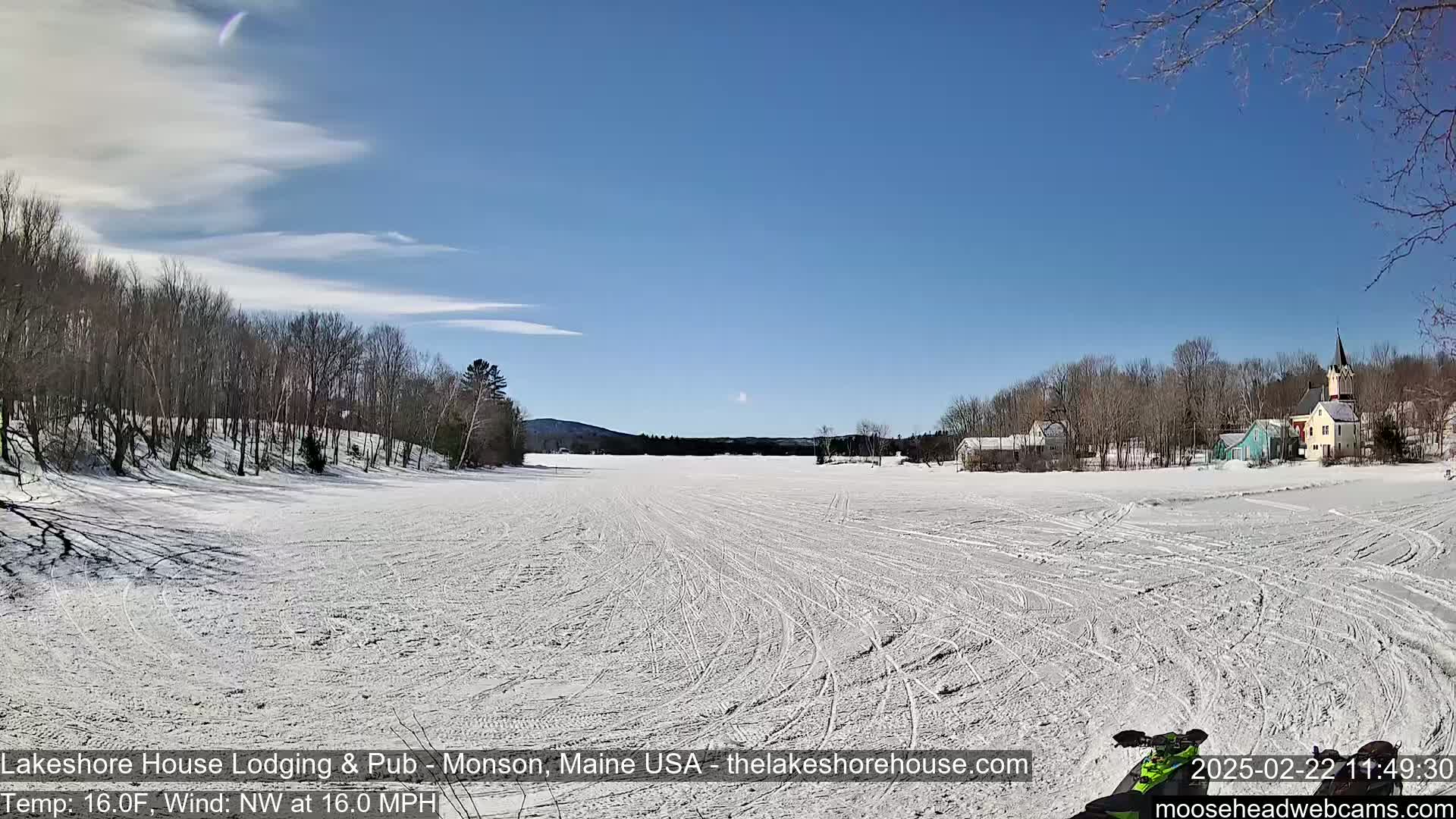 A snow-covered frozen lake stretches toward a small village of houses and a church under a clear blue sky with a light wind.